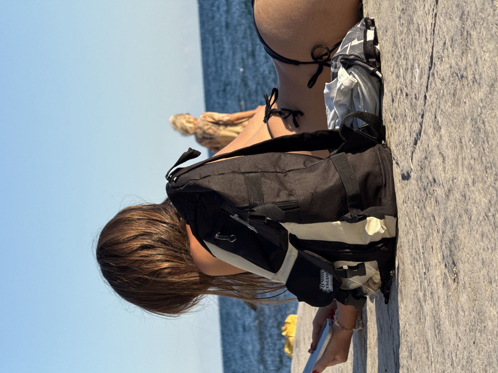 An outdoor shot shows a young woman lying on a stone surface with a black and cream backpack in the foreground. She is wearing a black bikini and is looking at her phone. The sea and sky are visible in the background, with another person walking in the distance. A yellow object is partially visible on the left side of the image. The scene is lit by sunlight.