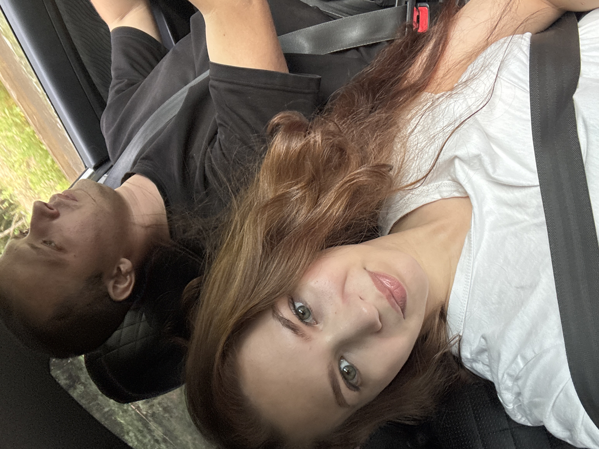 This media file shows a close-up, overhead shot of two people in a car, likely on a road trip. The subjects are lying down, with one person's head resting on the other's shoulder, suggesting intimacy or comfort. The person in the foreground is a woman with long, wavy brown hair and green eyes, wearing a white t-shirt. She is looking towards the camera with a slight smile and full lips. The person behind her is a man with short brown hair and a darker complexion, wearing a black t-shirt. He is looking away from the camera, with his eyes closed and his head tilted back, appearing relaxed or asleep. Both individuals are wearing seatbelts, indicating they are in a moving vehicle. The car's interior, including the black leather seats and seatbelt mechanisms, is visible. Outside the car window, blurred green foliage suggests a natural, possibly rural setting. The lighting is soft, implying it could be daytime, but the exact time is not discernible. There is no visible text in the image. The scene evokes a sense of casual travel and companionship. The crops highlight individual features like the woman's makeup and the man's profile, as well as details of their clothing and the car's interior.