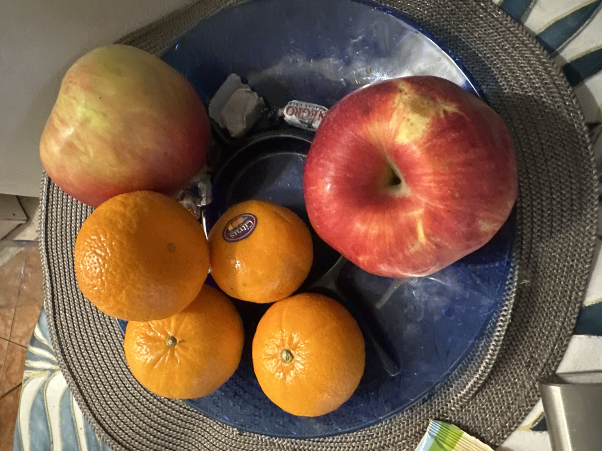 The image displays a close-up, overhead view of a still life composition on a tabletop in Budva, Montenegro. A circular, dark blue, textured plate contains five orange citrus fruits and two apples. One of the oranges bears a "Citrus" sticker. The apples exhibit varied coloring, ranging from predominantly red with yellow mottling on the lower apple, which features a distinct apical dimple, to a lighter red, yellow, and green blend on the upper apple. Positioned on the plate alongside the fruit are a small, unwrapped, light-colored candy piece and a candy wrapper with "Negro" text visible. A corner of a green, white, and blue striped wrapper is partially visible near the lower-left edge of the frame. The blue plate rests on a circular, woven placemat of dark grey material. This placemat is situated on a table surface characterized by an abstract pattern featuring white, light blue, and brown hues. A metallic object is partially observable in the lower-left foreground.