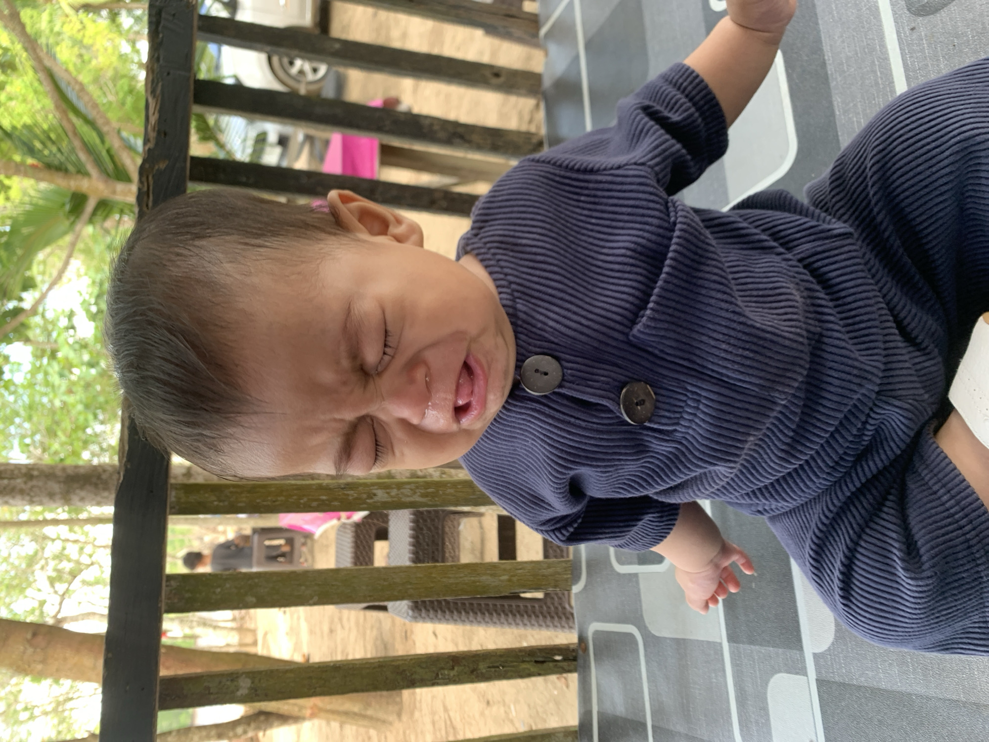 A baby in a navy blue, ribbed jumpsuit is crying, with eyes closed and tongue slightly out. The baby is lying on a gray and white patterned surface. Behind the baby is a wooden railing overlooking an outdoor area with trees and chairs. The location is Tanjung Limau, Indonesia.