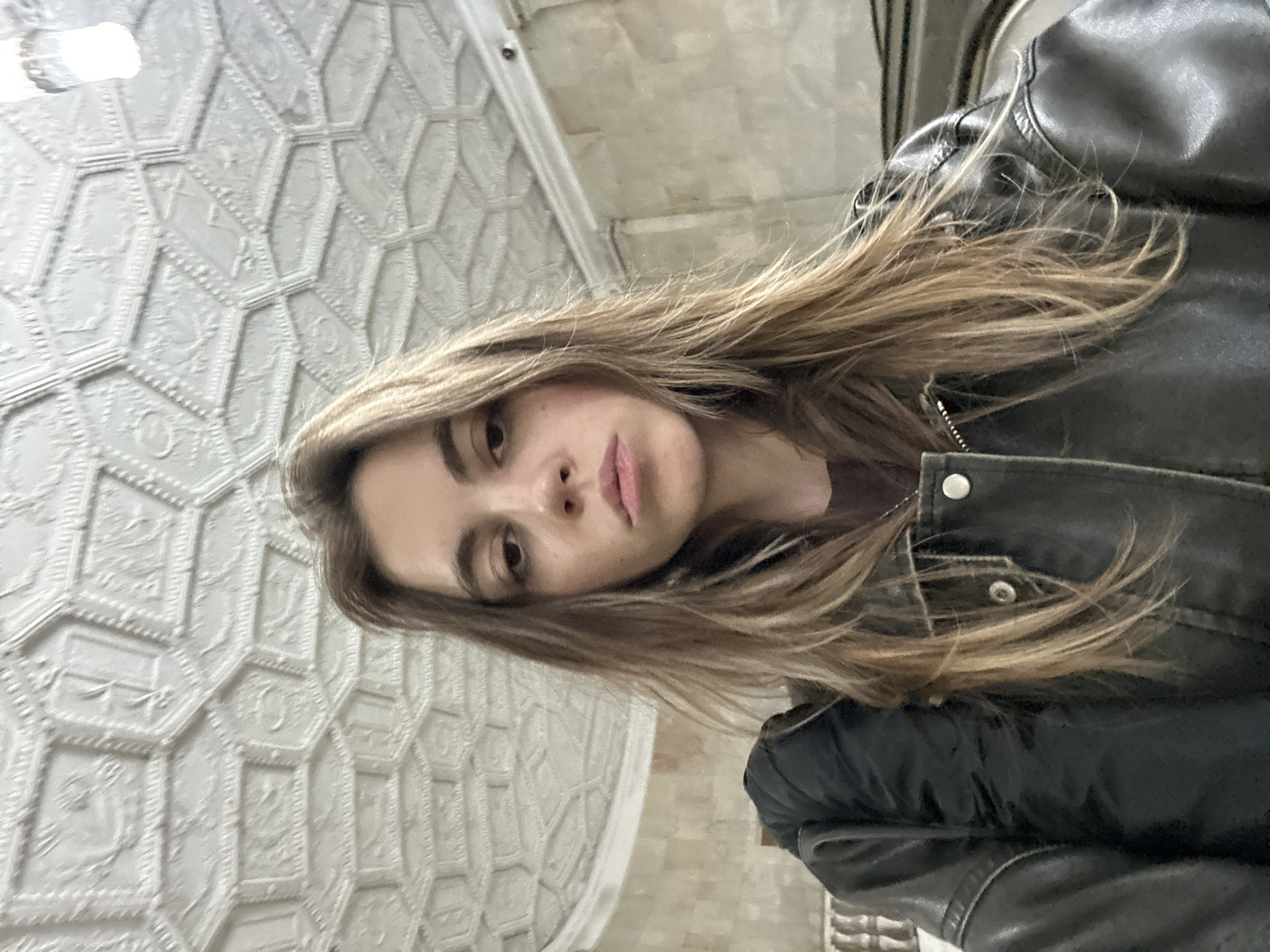 A young Caucasian woman with long, light brown hair is pictured from the chest up, looking directly at the camera. She is wearing a dark brown shirt and a black leather jacket with silver snaps. The background features a white, ornate ceiling with a honeycomb-like pattern, and walls made of textured beige stone. The overall setting appears to be an indoor public space, possibly a metro station, given the architectural style of the ceiling and the presence of what looks like an escalator railing in the background. The lighting is soft and diffused. There are no other people visible in the image. The woman's facial expression is neutral, with her lips slightly parted.