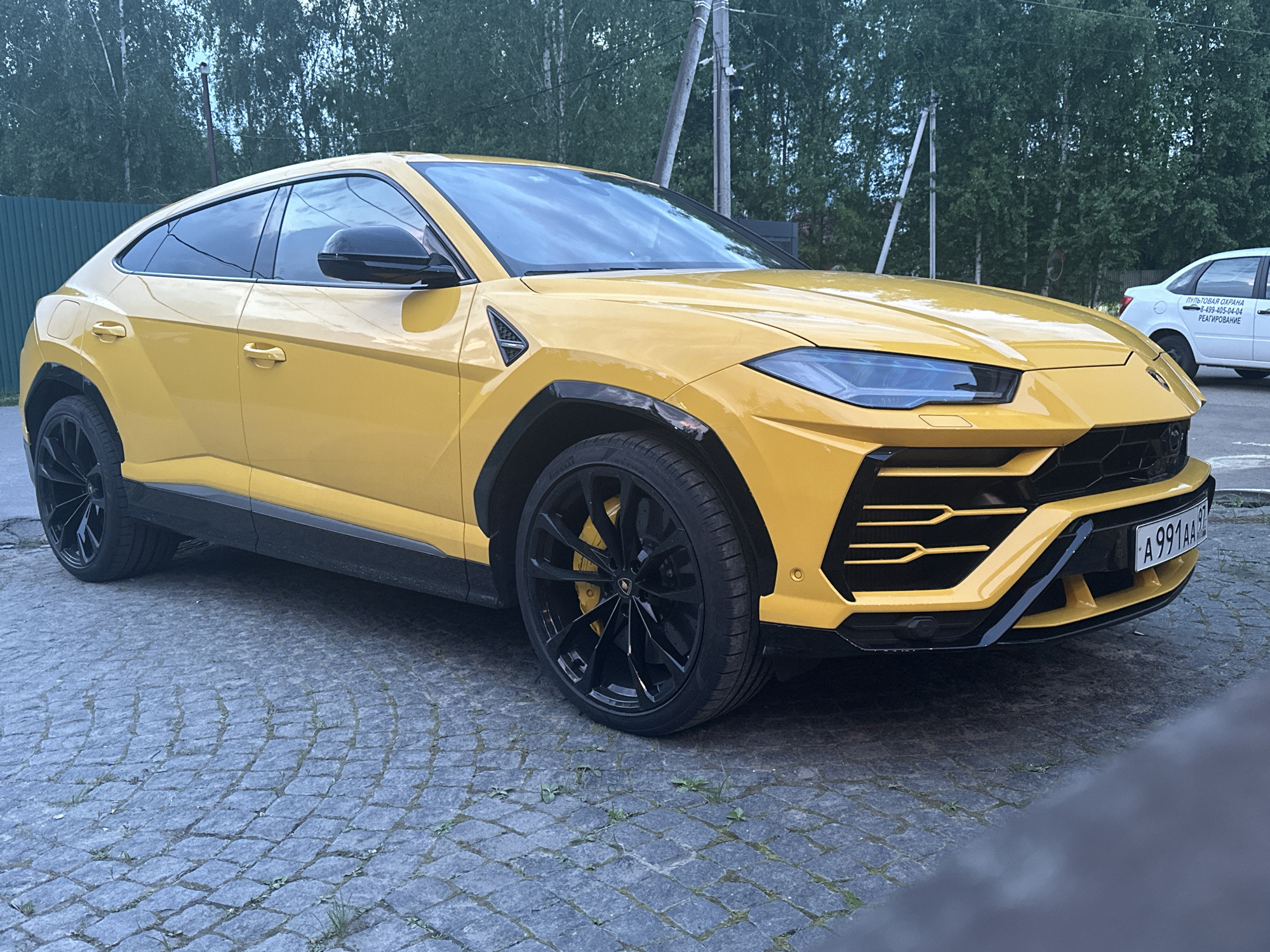 The image shows a yellow Lamborghini Urus parked on a cobblestone surface. The car has black wheels and accents. In the background, there is a green fence, trees, power lines, and a white car. The license plate on the Lamborghini is "A991AA97". The location is Новоглаголево, Russia, based on the license plate region code.