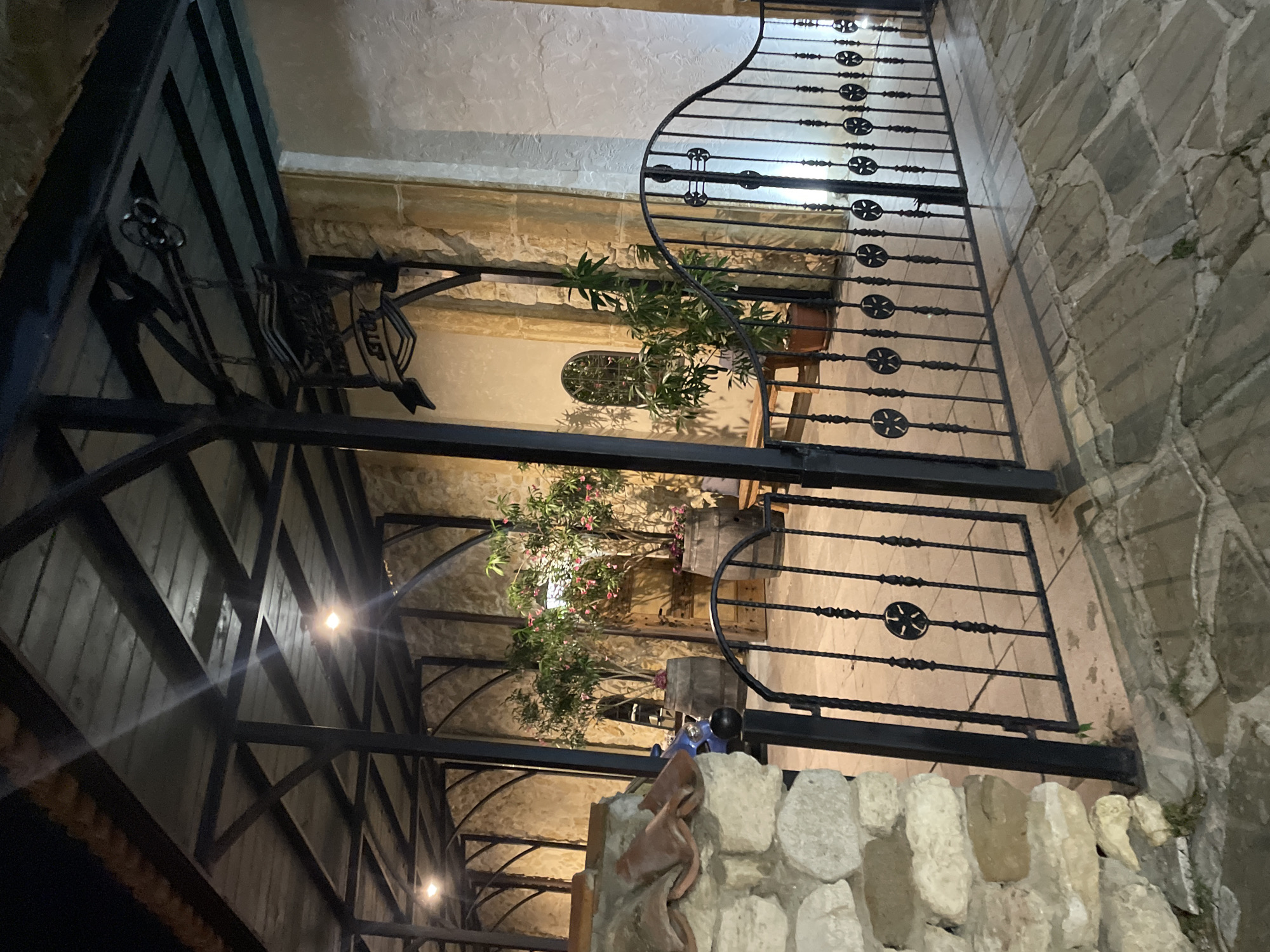 The image captures an outdoor patio area at night, likely a restaurant or a hotel in Nesebar, Bulgaria. The scene is illuminated by artificial lights. A black wrought-iron gate with decorative circular motifs is prominently featured in the foreground, opening onto a paved stone walkway. Beyond the gate, a covered walkway with a wooden ceiling and dark metal beams provides shelter. Two large wooden barrels are used as planters for trees, one with small pink flowers, situated in the covered area. A stone wall constructed from irregular-shaped stones is visible on the left. A sign with text that appears to read "HOTEL" is hanging from the ceiling of the covered area. The overall impression is one of rustic charm and a warm, inviting atmosphere. There are no people visible in the image.
