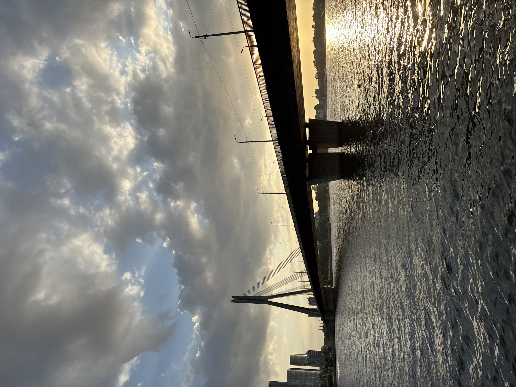 This is a photograph of the Vanšu Bridge in Riga, Latvia, taken at sunset. The cable-stayed bridge spans the Daugava River, connecting the city center with the Pārdaugava district. The image is dominated by the bridge itself, which is silhouetted against a dramatic sky filled with clouds. The sun is setting behind the bridge, casting a warm golden light on the water and creating a shimmering reflection.
In the background, on the left side of the image, are several tall modern buildings, characteristic of Riga's city skyline. On the right bank, a line of trees and some sailboats are visible, adding to the picturesque scene. The river in the foreground appears calm, with gentle ripples reflecting the light. Several streetlights are visible along the top of the bridge, and a few silhouetted figures can be seen walking on the pedestrian path. The overall impression is one of urban beauty and tranquility at the end of the day.