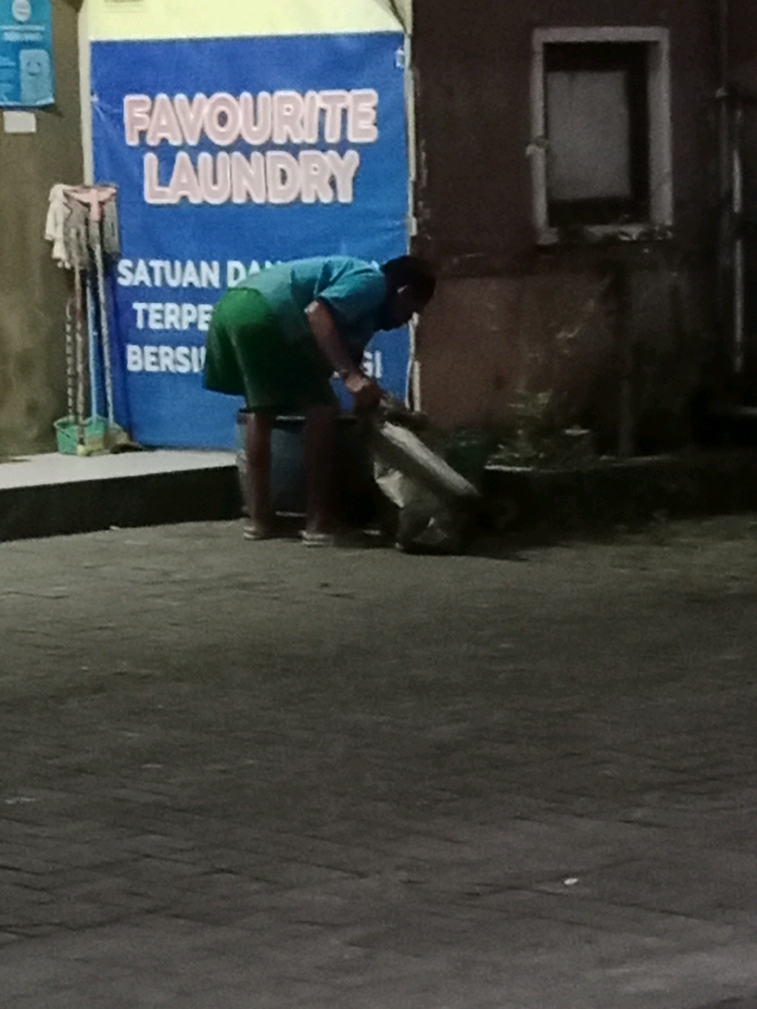 The image depicts an outdoor scene at night, likely in Waru, Indonesia, indicated by the text on a blue banner. A person wearing a teal shirt and green shorts is bent over, sweeping or gathering trash with a broom and a large piece of material, possibly a bag or cloth. The banner behind them displays "FAVOURITE LAUNDRY" in pink lettering, with additional text below that is partially obscured. To the left of the banner, a set of cleaning tools, including brooms and a green bucket, are visible. The ground is paved with dark-colored bricks. In the background, a dark, textured wall with a small, rectangular window can be seen. The overall lighting suggests it is nighttime or a dimly lit area.