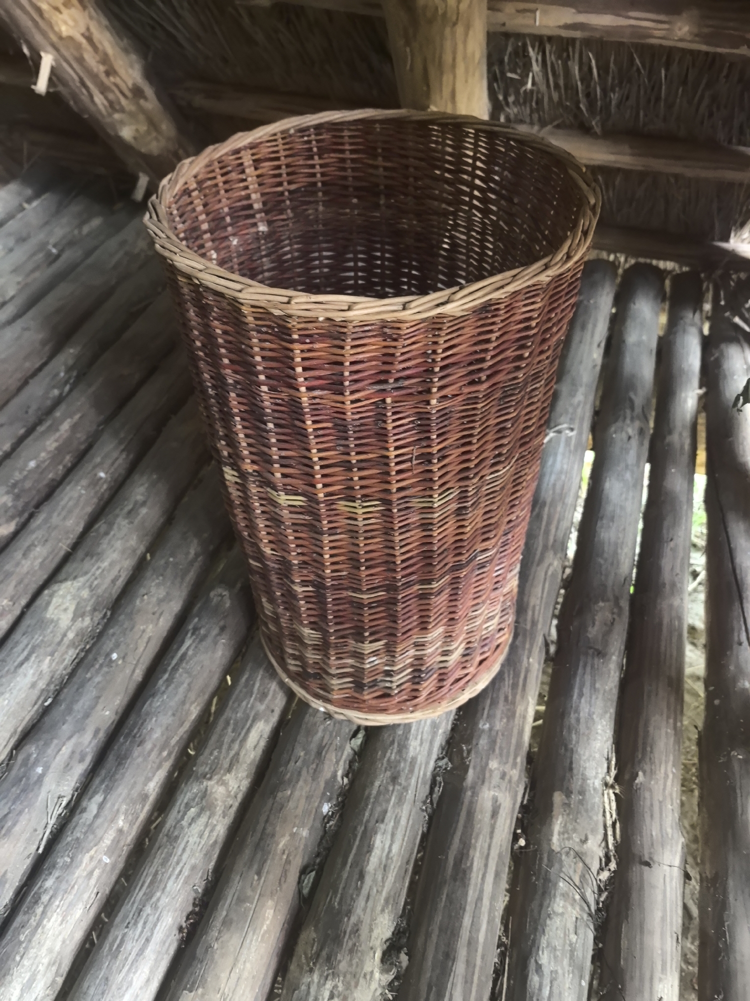 In the interior of a rustic structure, a wicker basket sits on a floor made of parallel wooden logs. The basket is woven from thin, flexible strands of a reddish-brown material, with a lighter colored rim. The logs are rough and weathered, with visible grains and occasional darker spots. Above the basket, the ceiling is constructed from similar wooden beams and a thatched material. The overall impression is one of natural materials and a simple, handmade construction, possibly in a rural or historical setting. The location of Камянюцкі сельскі Савет, Belarus, suggests this could be a traditional dwelling or outbuilding in a Belarusian village. No people are present in the image.
