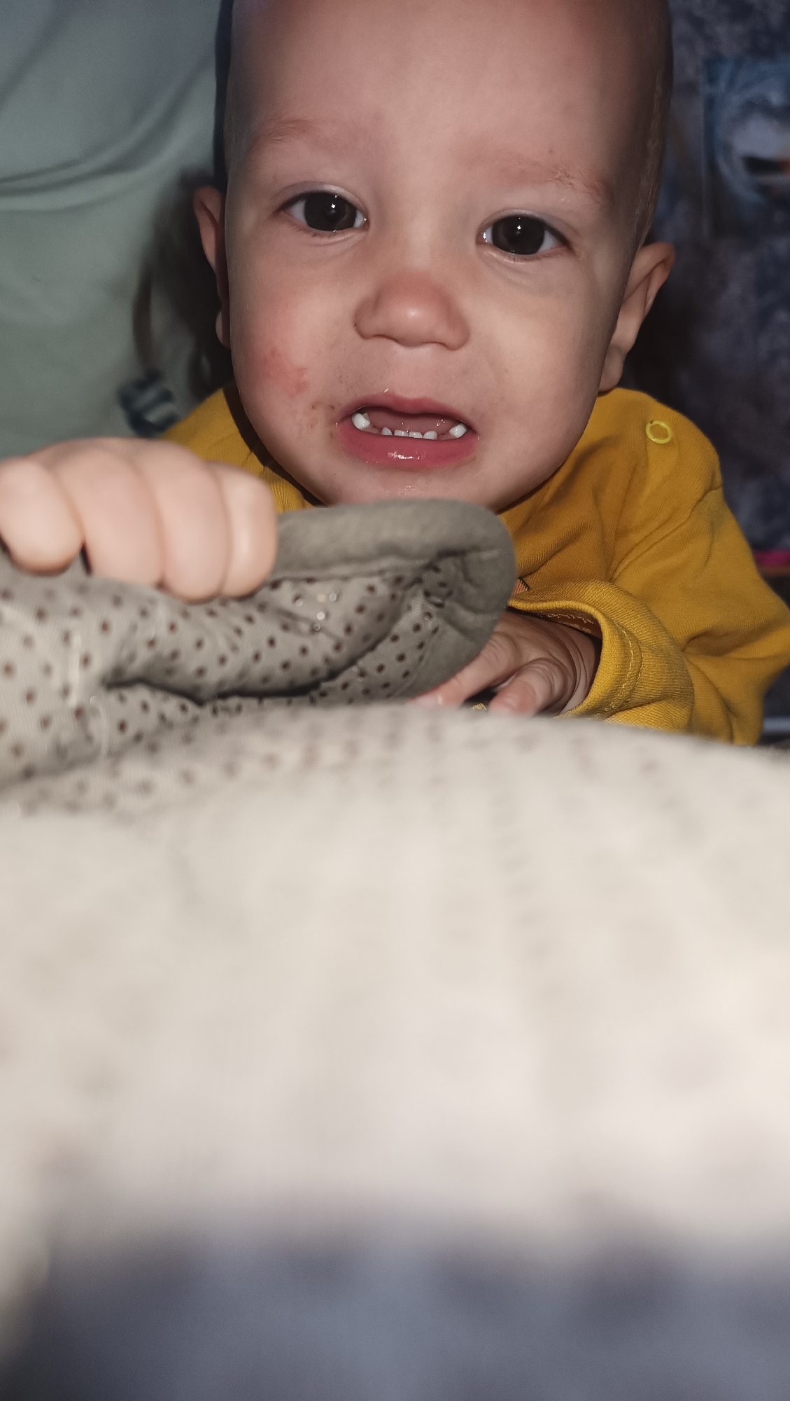 A close-up, low-angle shot shows a young child, appearing to be a toddler, with dark eyes and fair skin. The child is wearing a yellow long-sleeved shirt and is looking directly at the camera. The child's mouth is slightly open, revealing several small teeth. Red marks and some dark smudges are visible on the child's face, particularly around the mouth. The child's right hand is visible, resting on a gray, dotted blanket. In the foreground, a fuzzy, light-colored blanket or textile obscures the lower portion of the frame. The background is blurred, with hints of other fabrics and possibly a wall. The image does not contain any identifiable landmarks or details that would indicate its location in Karsha, Russia, or any other specific city.