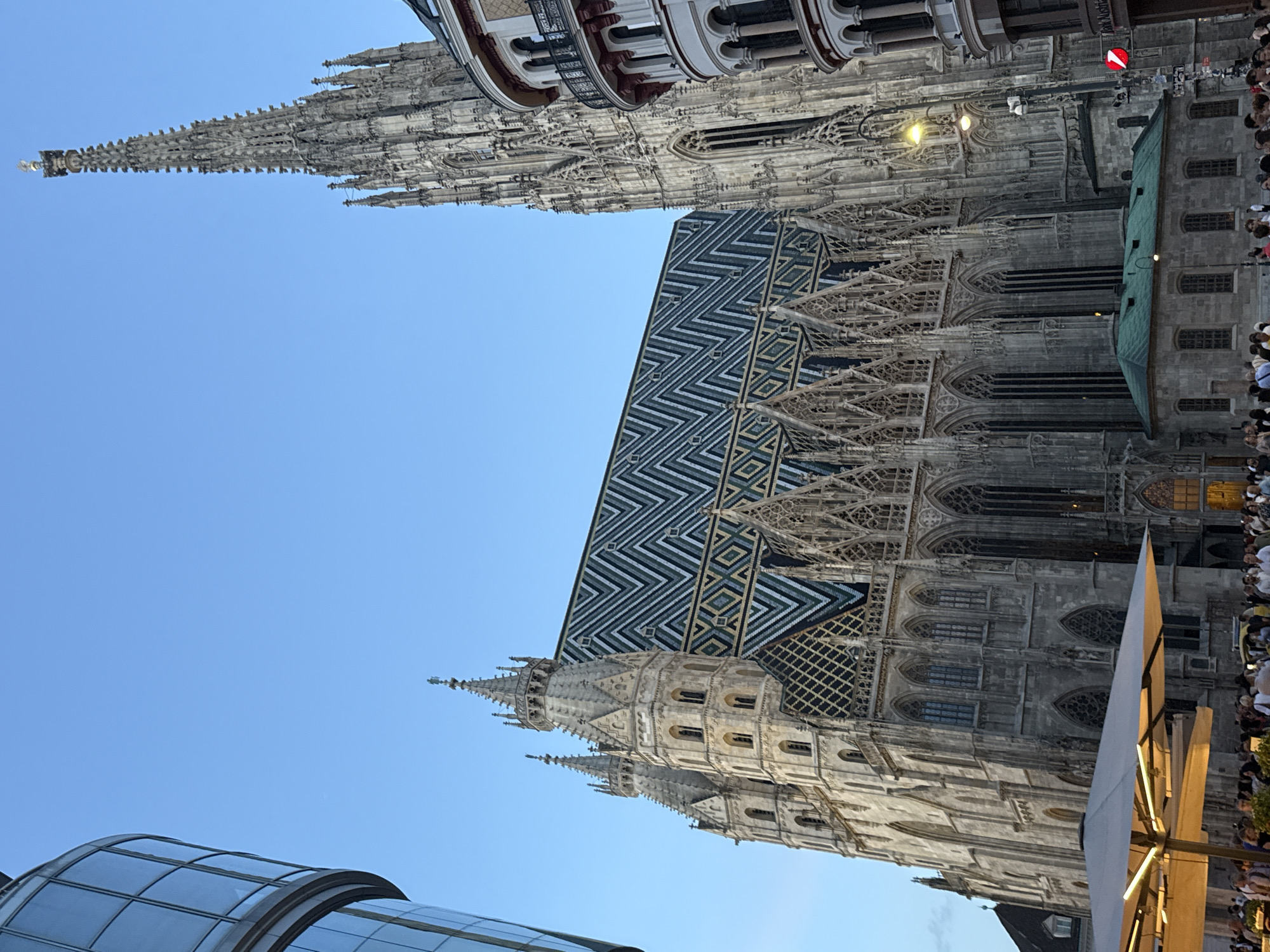 This media file depicts the St. Stephen's Cathedral in Vienna, Austria. The image showcases the intricate Gothic architecture of the cathedral, with a focus on its spires and a vividly patterned tiled roof. A modern glass structure is visible in the foreground, contrasting with the historical edifice. A crowd of people can be seen on the right side of the image, suggesting a public gathering or tourist activity. The sky is clear and blue, indicating fair weather and likely daytime, possibly late afternoon given the lighting. The overall scene is one of historical grandeur set against a contemporary urban backdrop, with a sense of bustling activity from the onlookers. No visible text is present in the image.
