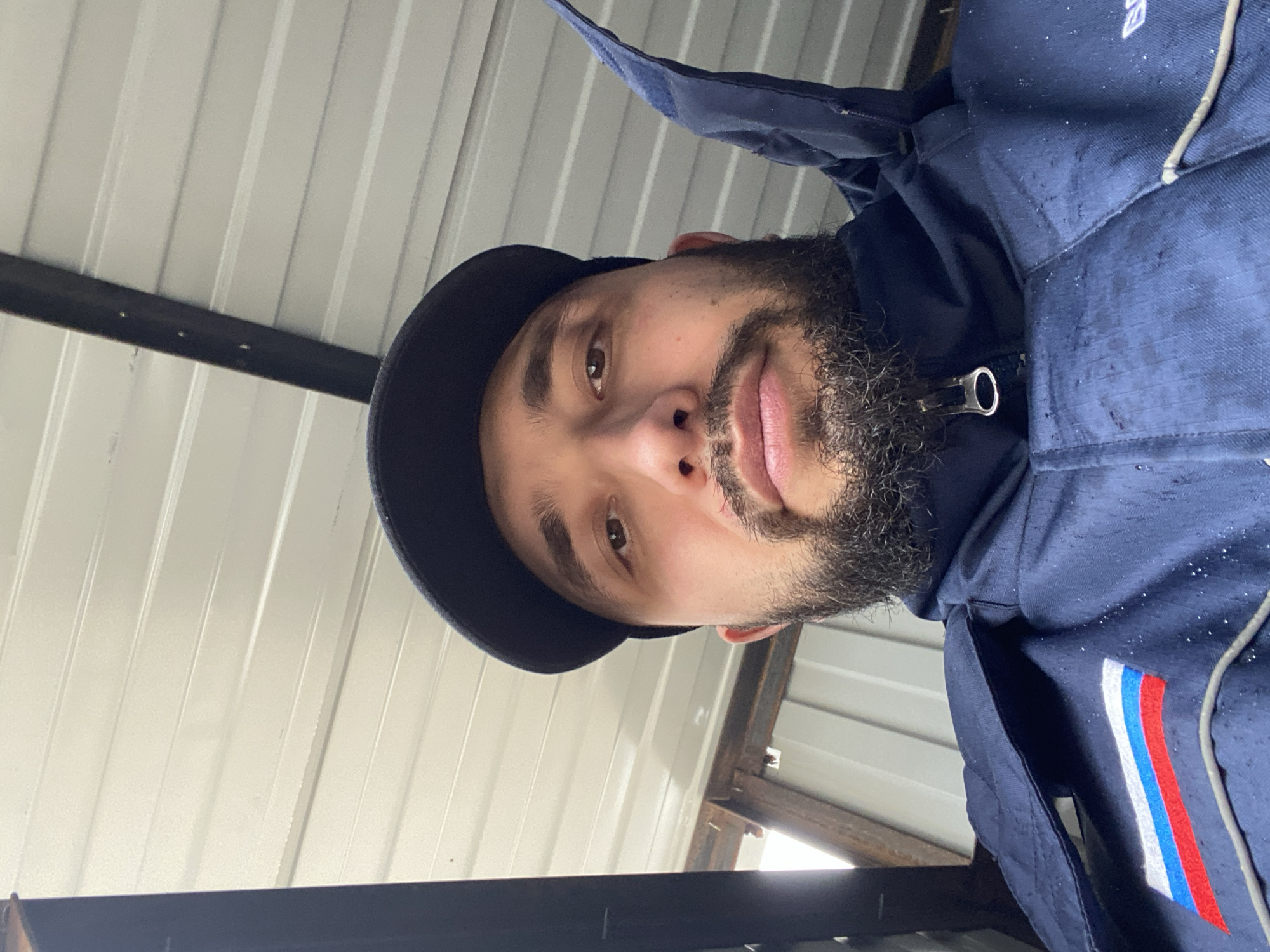 A man with a beard is facing the camera. He is wearing a dark cap and a blue jacket with a red, white, and blue stripe on the right side. The background is a corrugated metal ceiling with visible framing. The location is not identifiable as Luzhitsy, Russia based on the given visual information.