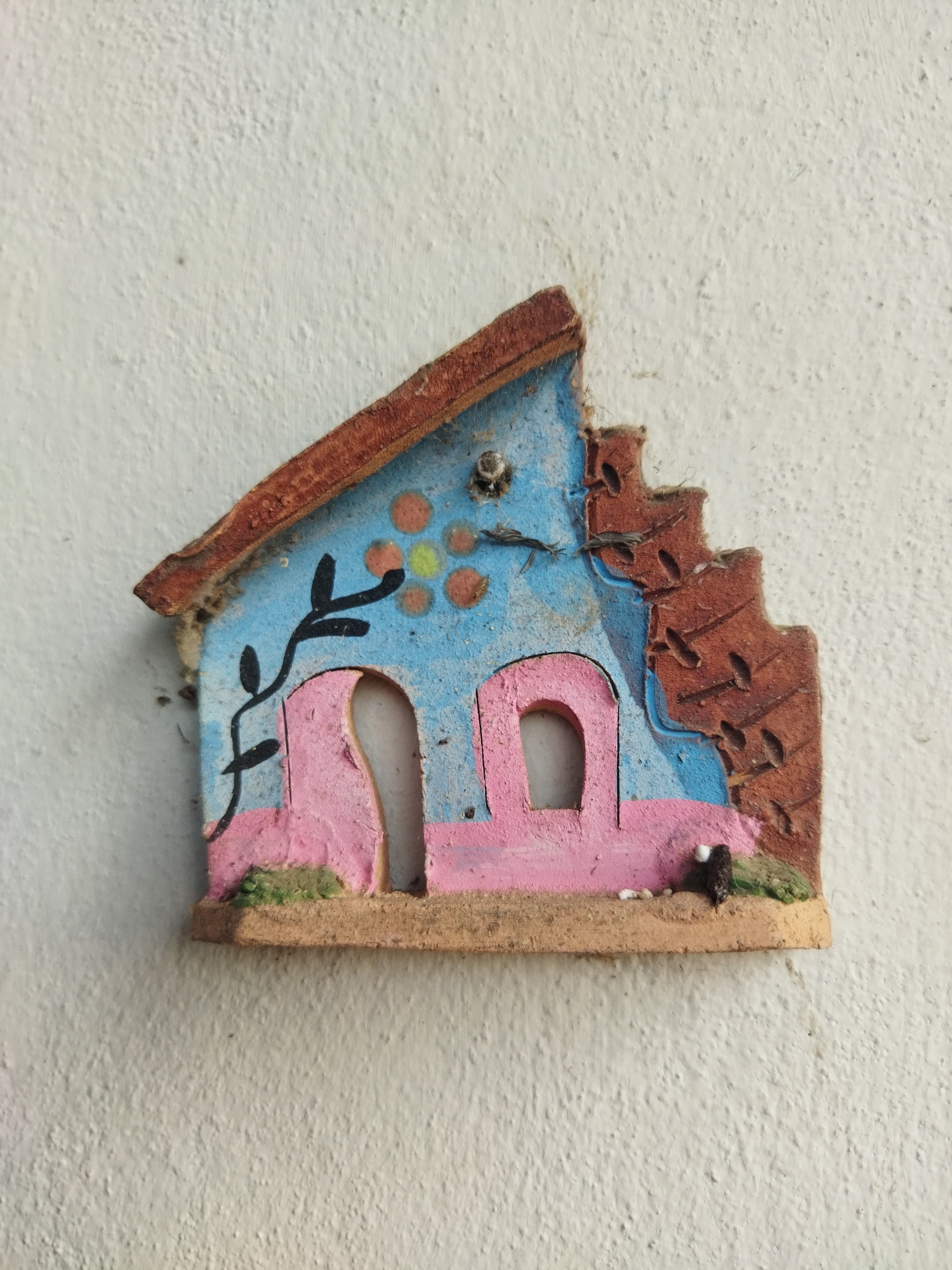 A small, colorful, hand-painted ceramic house is affixed to a white, textured wall. The house is predominantly blue and pink, with a brown roof and base. The roof is slightly damaged on the right side, revealing a brick-like pattern. The house has two windows and a door, all painted pink. A black painted vine climbs the left side of the house, and a cluster of orange and yellow circles are near the top. The base of the house is a sandy color with small details suggesting vegetation and debris. The image was taken in the city of Tinaquillo, Venezuela.