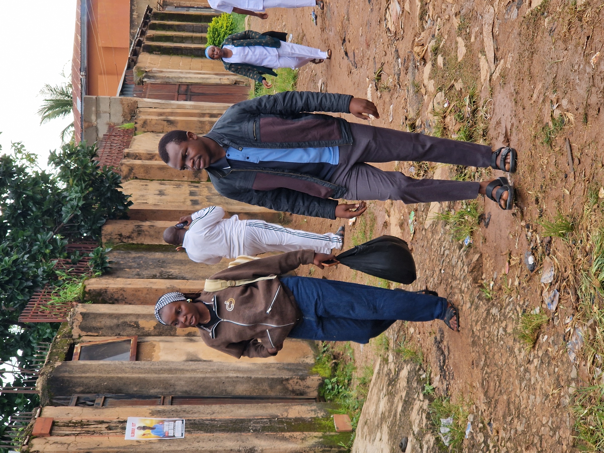 A man and a woman are walking down a dirt road in Jos, Nigeria. The woman is in front, carrying a black bag in her right hand. She is wearing a brown jacket, blue jeans, and sandals. The man is behind her, wearing a dark jacket over a blue shirt and gray pants, and sandals. A man in a white shirt and pants is walking in the background, talking on his phone. Another woman in white is walking further back. The road is unpaved and littered with debris. A concrete wall with a metal fence runs along the left side of the road, with a poster on it. The scene appears to be in a residential area.