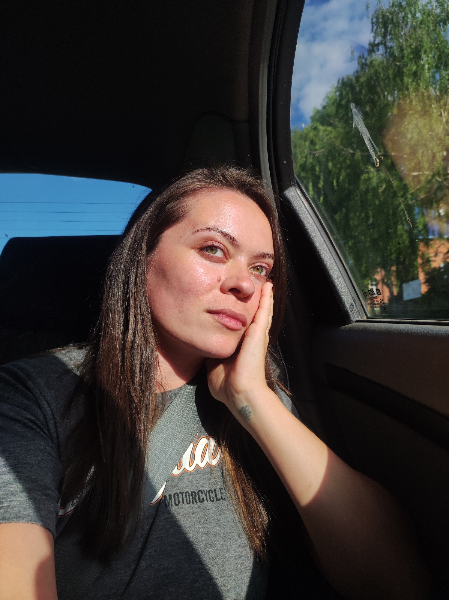 A woman with long, brown hair and light skin is sitting in the passenger seat of a car. She is looking out the window, her right hand resting on her cheek. Sunlight streams into the car, illuminating her face and casting shadows. She is wearing a gray t-shirt with the word "MOTORCYCLE" printed on it in white and orange lettering. The car's interior is visible, including a dark-colored seat and the window frame. Outside the window, trees and a blue sky with some clouds are visible. The overall impression is one of contemplation or quiet observation. The location is identified as the city of Fokino, Russia.