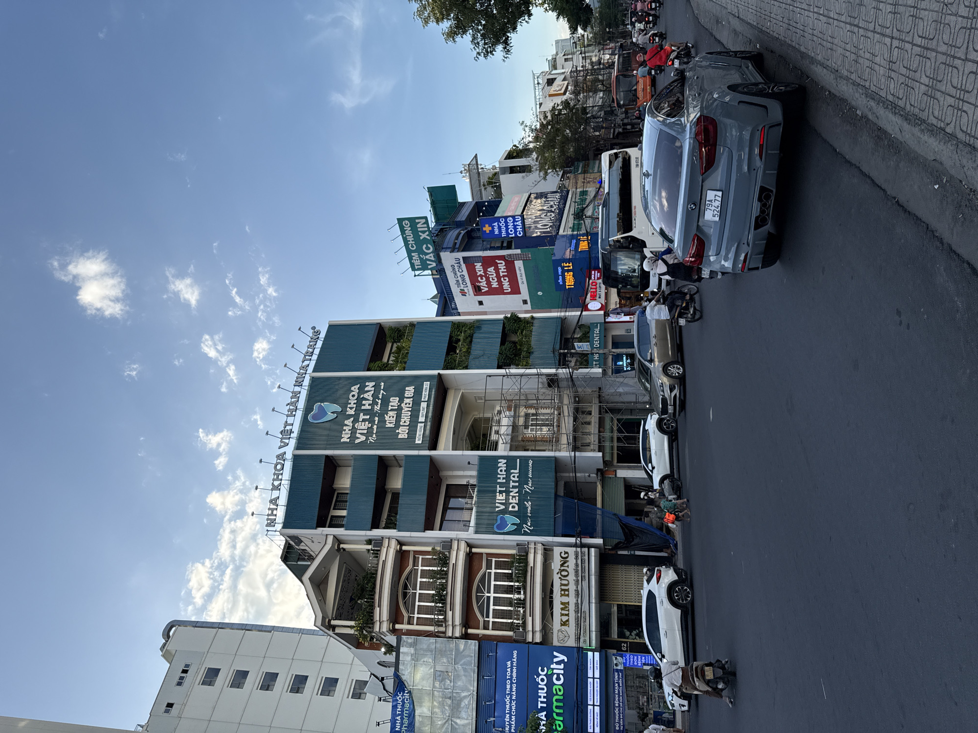 The image depicts a street scene in Nha Trang, Vietnam, under a clear blue sky with scattered clouds. The foreground is dominated by a gray BMW X6 parked on the right side of the road, its rear facing the camera. The car has a license plate reading "79A 524 77". To the left of the car, the road is paved with asphalt, and a patterned sidewalk runs along the right edge.
In the background, a busy street unfolds with various buildings and vehicles. Prominent on the left is a tall, white building with multiple stories and windows. Adjacent to it is a teal and white building with signage for "NHA KHOA VIET HAN DENTAL" (Viet Han Dental Clinic) displayed prominently. This building also features several floors with balconies and greenery. Other businesses visible include "PHARMACIT", "KIM HUONG", and "TIEM CHUNG VACCINE", indicating a commercial area with various services.
Traffic on the street includes several white SUVs, a white bus, and numerous motorcycles, some carrying one or two riders. A person on a motorcycle is in the immediate foreground on the left, heading away from the camera. Two children are walking on the sidewalk in front of the teal and white building. The overall impression is of an active urban environment in Vietnam.