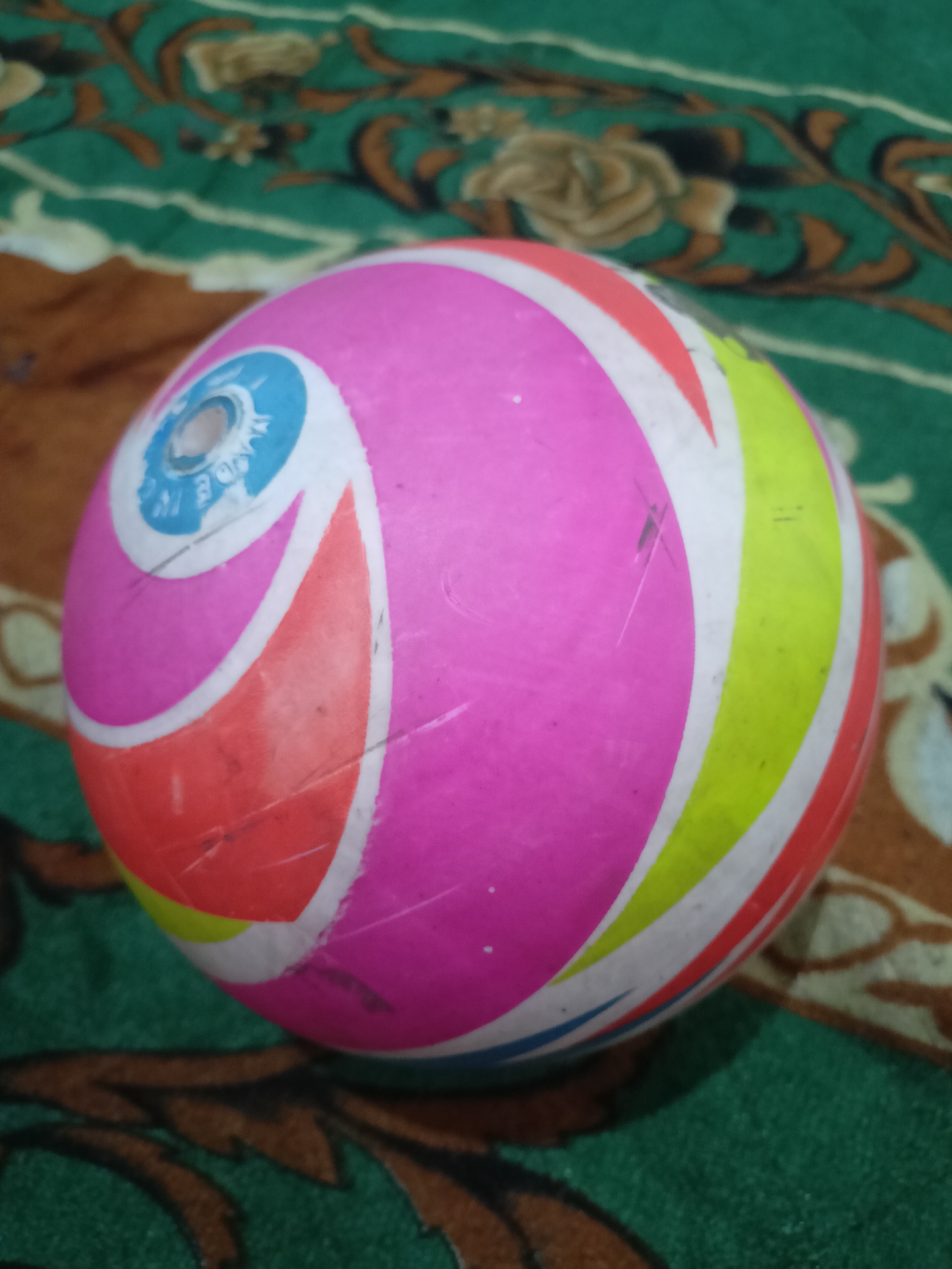 A colorful, striped beach ball rests on a patterned green and brown rug. The ball is primarily pink with curved stripes of white, orange, yellow, and blue. A small circular indentation is visible on the upper left portion of the ball. The rug has a floral or leaf-like pattern in dark green and brown. The lighting is dim, casting a slight shadow on the rug. The image is taken from a slightly elevated, close-up perspective.
