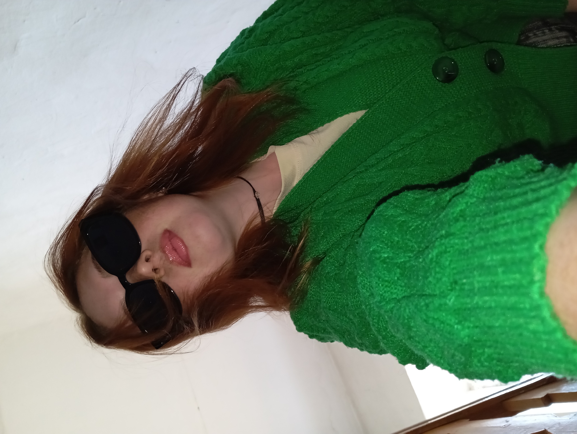 A woman with red hair and black sunglasses poses in front of a white wall, wearing a green knitted cardigan over a cream-colored shirt. The location cannot be verified as Yurgamysh, Russia, based on the provided image.