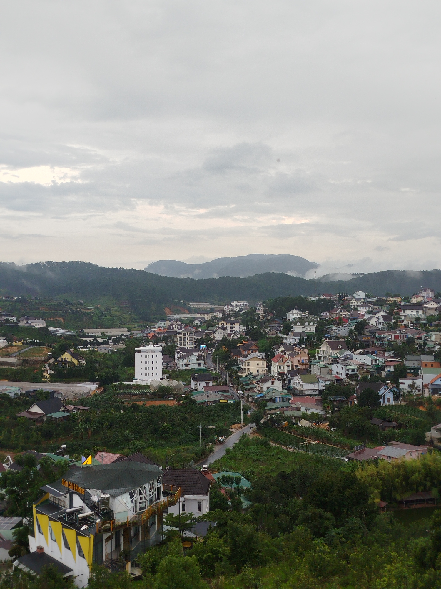 This is a high-angle, wide shot of a densely populated hillside town in Da Lat, Vietnam, under a cloudy sky. The scene depicts a multitude of buildings, varying in size and architectural style, nestled amidst lush green vegetation and rolling hills. In the foreground, a prominent multi-story building with a yellow and white facade and a dark roof stands out, featuring angular window designs and what appears to be an outdoor seating area. Further into the town, a tall, white, rectangular building with numerous windows is visible, along with a network of roads winding through the houses. The surrounding landscape is characterized by steep slopes covered in trees and shrubs, with some areas showing evidence of agricultural terraces or greenhouses. The overall impression is one of a vibrant, somewhat dense urban environment integrated into a hilly, natural landscape.