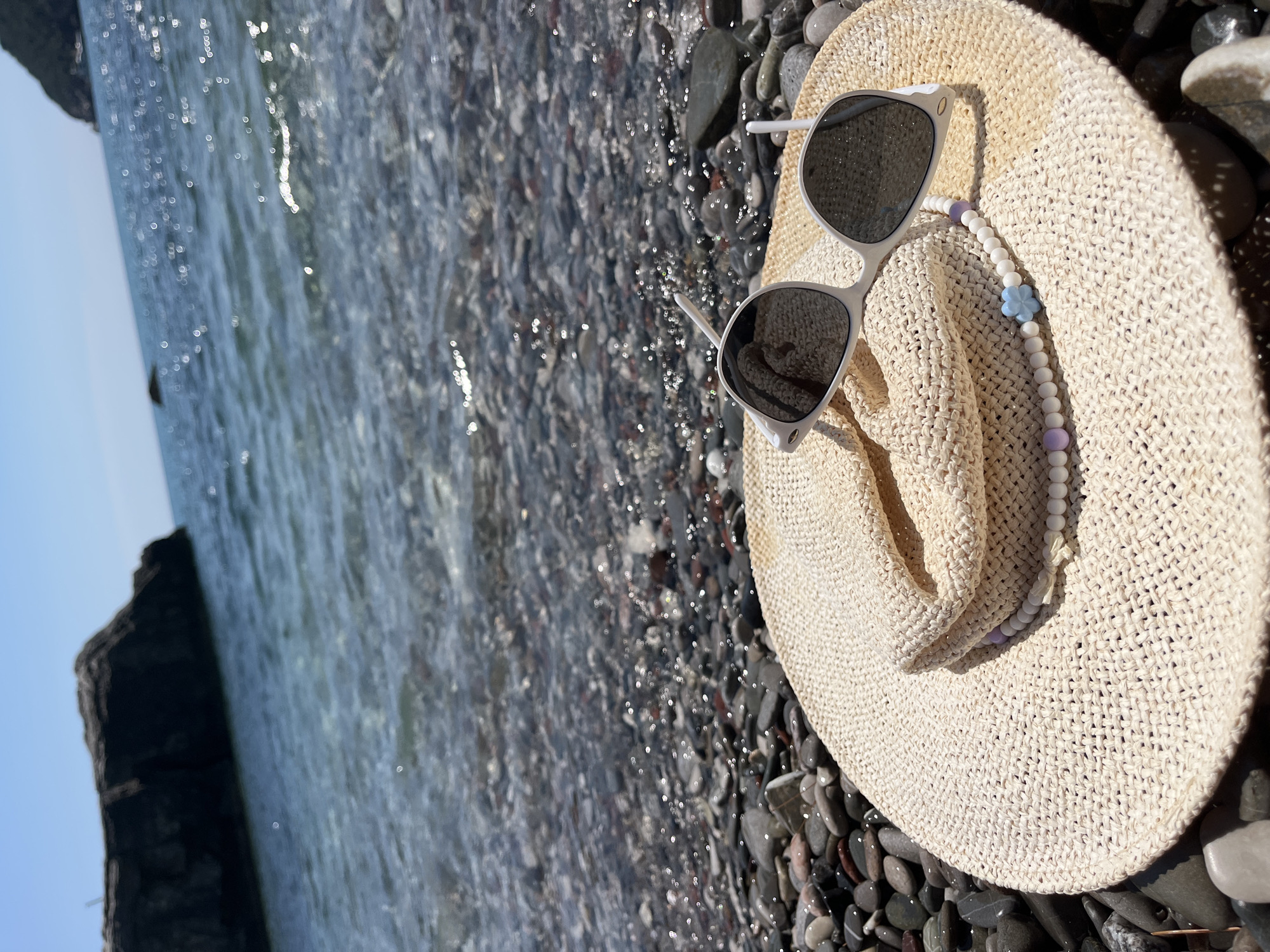 In Zagrađe, Montenegro, a straw hat and sunglasses lie on a pebble beach. The beach is adjacent to the clear, blue water of the Adriatic Sea, which reflects the bright sunlight. In the background, a dark, rocky outcrop juts out from the sea. The hat is light beige and has a decorative string of white and pastel beads with a blue flower charm. The sunglasses are white with dark lenses. The scene depicts a serene beach setting on a sunny day.