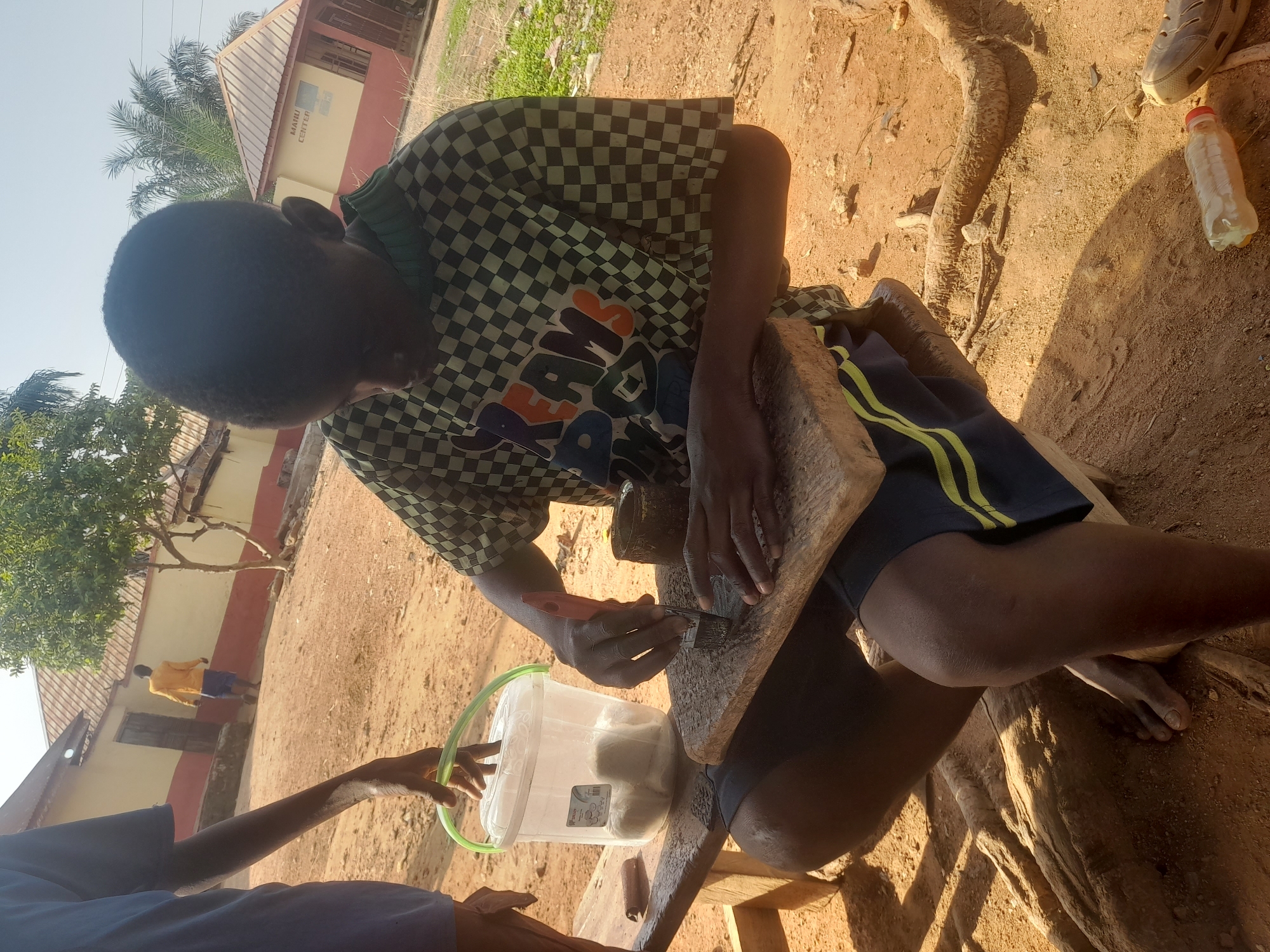 The image shows a person sitting on a wooden stool in an outdoor environment, potentially in Akwanga, Nigeria. The individual is wearing a checkered shirt and shorts with yellow stripes. They are holding a small tool and working on a flat piece of wood. A bucket containing white objects is positioned beside them. Another person's arm is visible, partially extending towards the bucket. The ground appears to be sandy and unpaved. In the background, there is a building with a sign reading "MANU CENTER...". A few other people and trees are also visible. A water bottle sits near the person's feet. The scene appears to be a local or rural setting, possibly involving craftsmanship or a manual task.