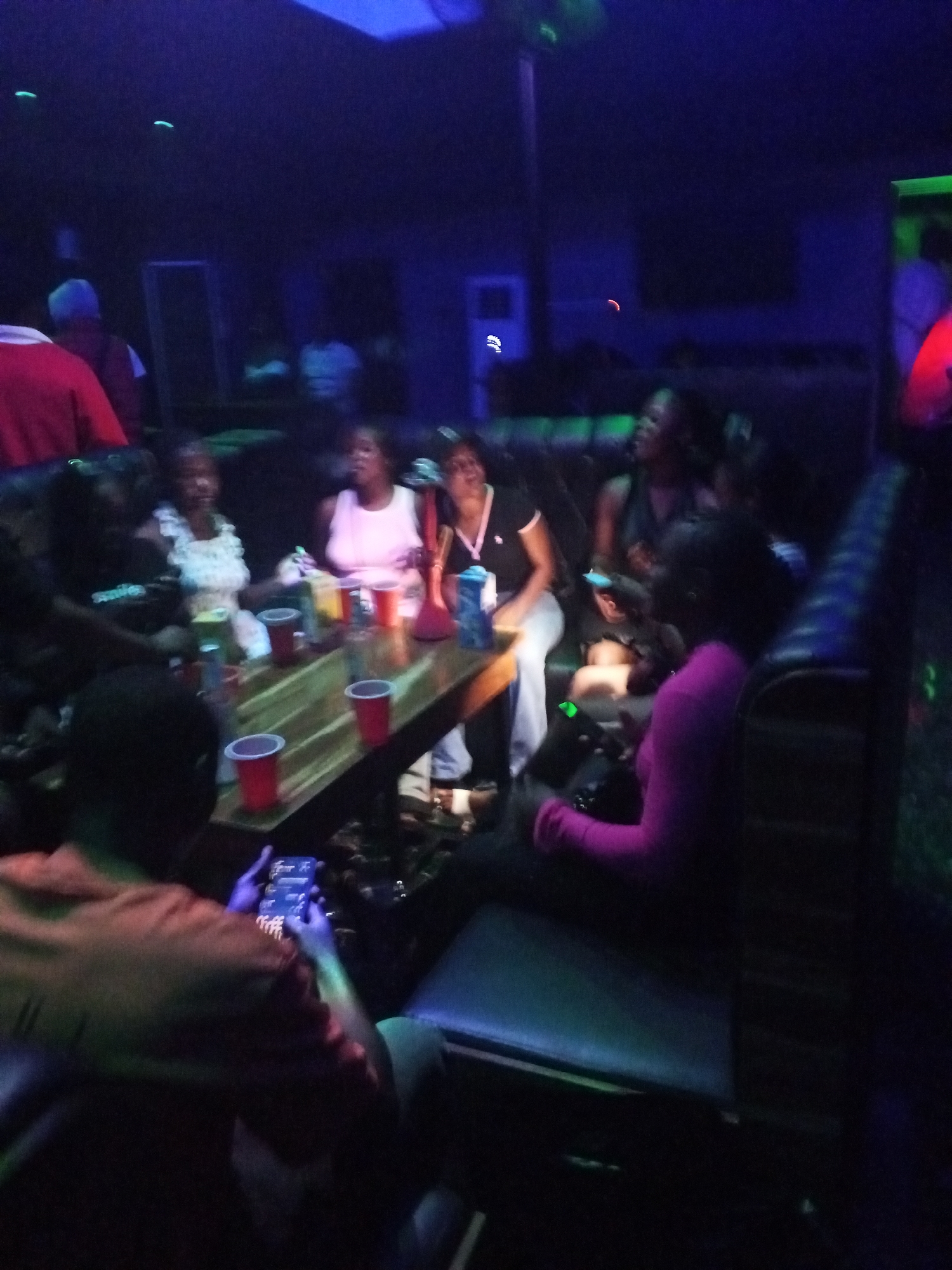 In a dimly lit indoor establishment, possibly a bar or lounge in Lokoja, Nigeria, a group of people are gathered around a wooden table. Red plastic cups and various drink containers are scattered on the table. A hookah is also present. The individuals, mostly adults, are dressed casually. Several are looking at or using their mobile phones. The room has a dark ambiance with blue and green lighting. The scene appears social and relaxed.