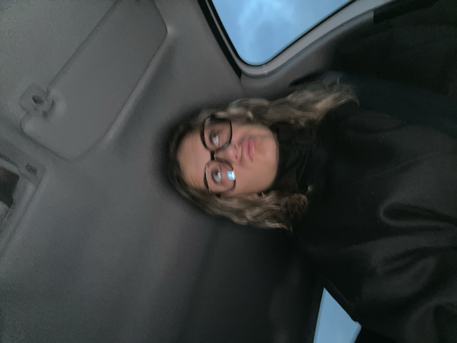 A young woman with curly blonde hair and large-framed glasses is inside a car. She wears a black top and is looking at the camera with a slightly pursed lip expression. The interior of the car is visible, including the sun visor and the window showing a cloudy sky. The perspective is from a low angle, making the roof and top of the car prominent in the frame.