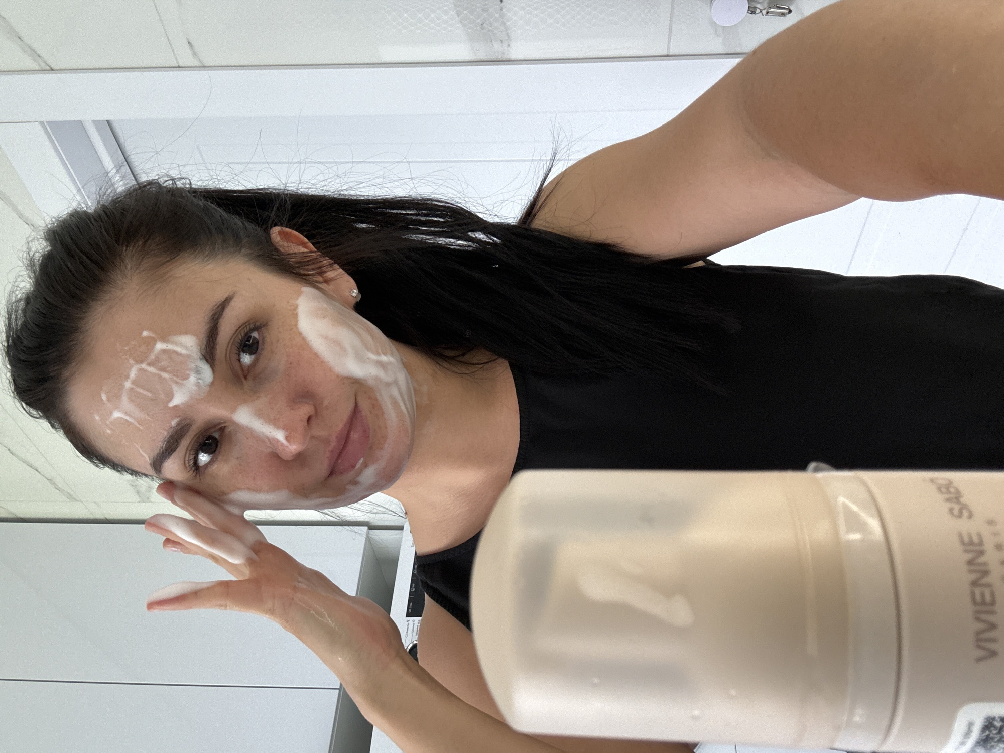 A woman with dark hair pulled back into a ponytail is applying foam cleanser to her face. She is wearing a black sleeveless top. Her face is partially covered in white foam, and she is holding her left hand to her cheek, gently massaging the cleanser. In the foreground, a beige bottle with a white cap is visible. The background appears to be a bathroom with white tiled walls and light gray cabinets. The scene is well-lit, suggesting it was taken indoors during the day. There is no indication of the location in the city of Нижнеяркеево, Russia.