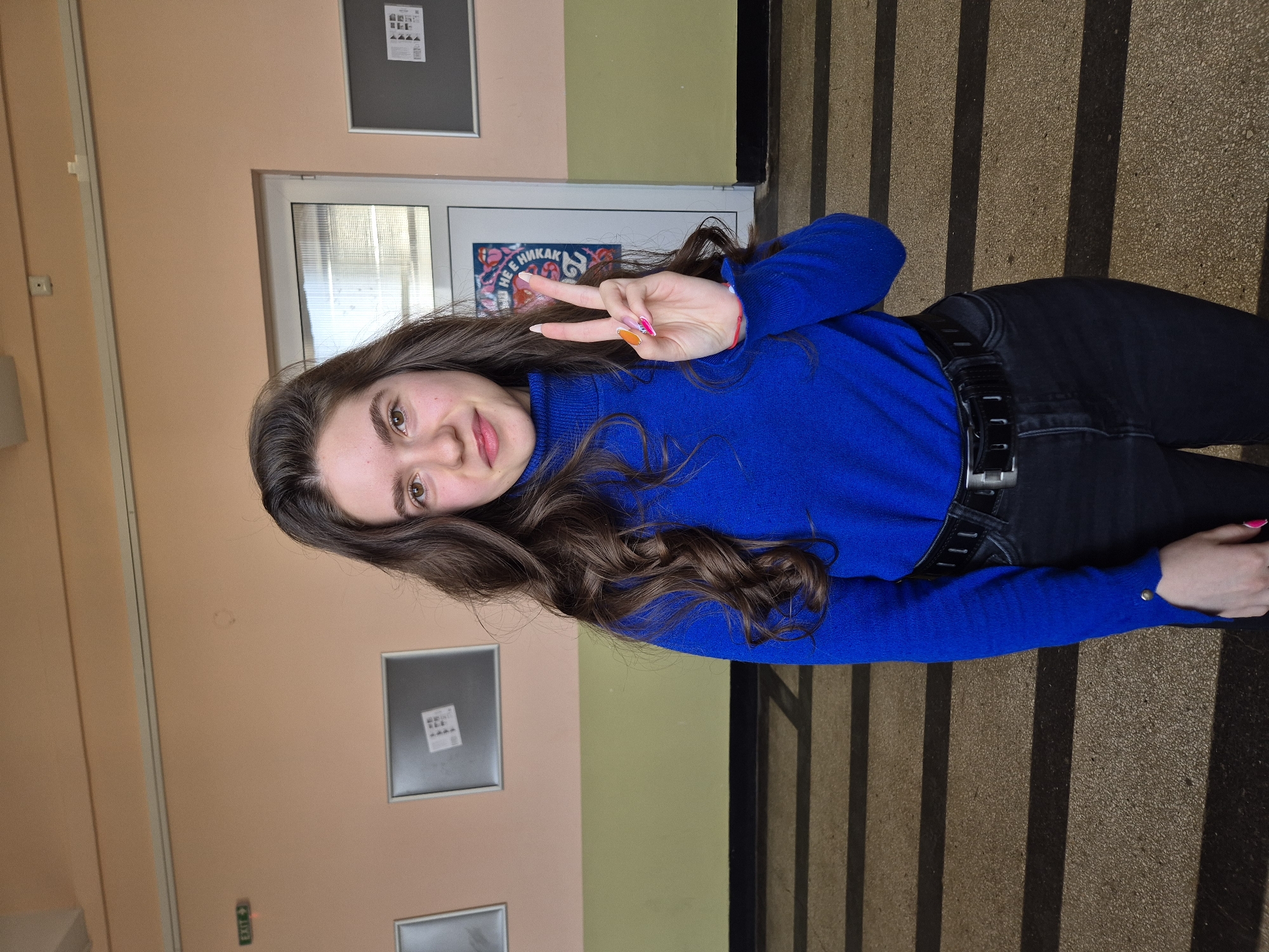 A young woman stands indoors, smiling and holding up a peace sign. She has long, wavy brown hair, hazel eyes, and light skin. She is wearing a bright blue turtleneck sweater, black jeans, and a black belt. The background features an interior wall with peach and green paneling. A doorway is behind her, with a sign visible in Bulgarian that says "НЕ Е НИКАК". The location appears to be a school in Pavlikeni, Bulgaria. The floor appears to be textured concrete with shadows that suggest a grated walkway or stairwell. There are notice boards on the wall.