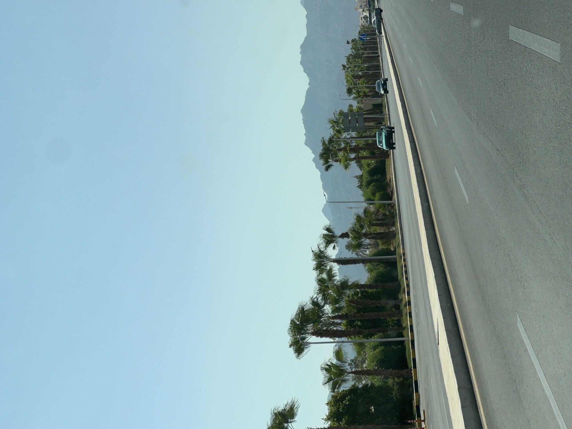 This is a medium-distance, eye-level, horizontal shot of a road in Sharm El Sheikh, Egypt. The road is paved and has lane markings. Several cars are driving on the road. On the left side of the road, there is a sidewalk with palm trees and other greenery behind it. In the background, there are mountains. The sky is clear and blue. The overall scene suggests a sunny day in a city.