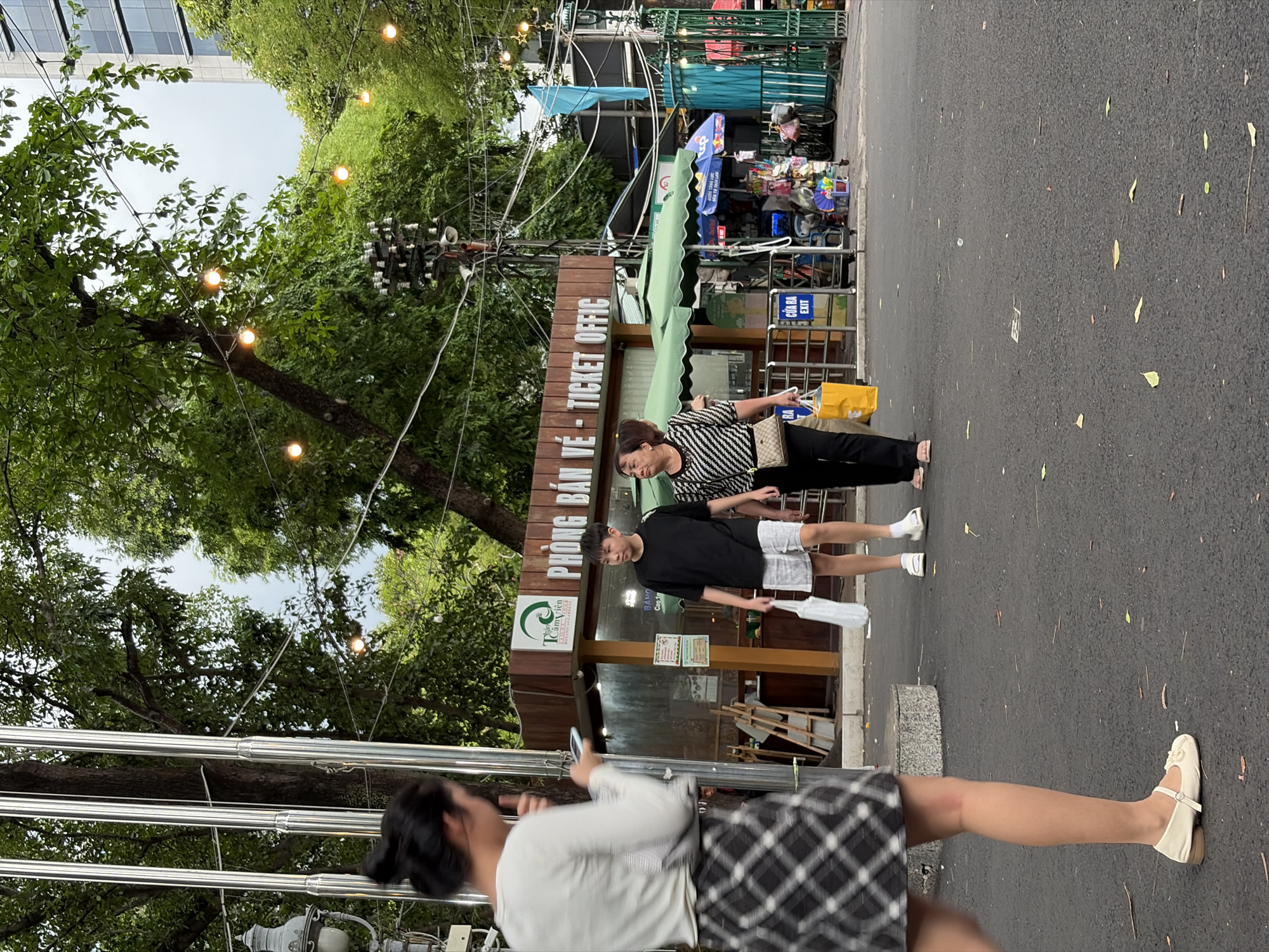 The image depicts an outdoor street scene at the entrance or exit of Thảo Cầm Viên Saigon Botanical Garden (Saigon Zoo and Botanical Garden) in Ho Chi Minh City, Vietnam. This area is part of the larger Ho Chi Minh City metropolitan region, which encompasses Thủ Đức.
In the foreground on the left, a young Asian woman is partially visible from behind, in mid-stride. Her body is blurred, indicating movement. She has dark hair styled in a bun and wears a long-sleeved white top and a black-and-white plaid skirt. She has light-colored, strapped flat shoes on her feet and is holding a smartphone in her left hand. To her left, several tall, reflective metallic poles ascend vertically.
In the mid-ground, slightly to the right of the center, an Asian young man and an older Asian woman are walking side-by-side towards the right. The young man, wearing a black t-shirt and white shorts, carries a white plastic bag in his left hand. The older woman, dressed in a black and white horizontally striped top and dark pants, carries a light-colored, patterned shoulder bag and a yellow shopping bag in her right hand.
Behind them, a single-story brown building with a green roof overhang serves as a ticket office, indicated by a sign reading "PHÒNG BÁN VÉ - TICKET OFFIC" and "Thảo Cầm Viên Saigon Botanical Garden" with a green leaf logo. To the right of the ticket office, several green market umbrellas shade small kiosks selling various items like fans, toys, and packaged goods. An "CỬA RA EXIT" sign is visible near a metallic barrier. A bicycle is parked near the stalls, and a blue flag hangs further in the background.
The upper portion of the scene is dominated by dense green foliage from numerous large trees. Warm-toned string lights are strung between these trees, creating an overhead canopy. A complex electrical utility pole with multiple wires and insulators is visible among the trees on the right side. The ground is dark asphalt pavement, sparsely scattered with light-colored leaves and small debris. An ornate street lamp is partially visible on the far left. The sky is overcast.