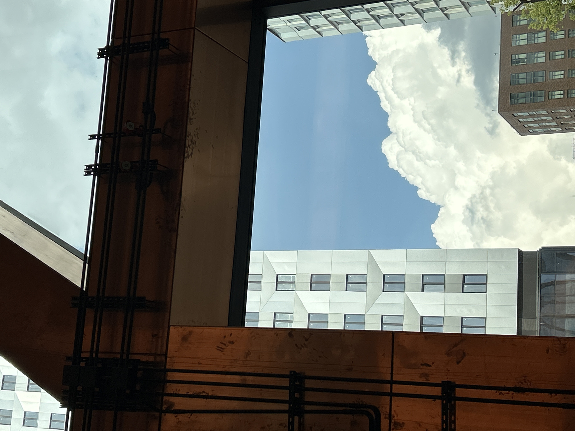 The photograph captures a view from an interior space, looking outwards through a large window or opening. In the foreground, a portion of the interior is visible, featuring a wooden structural element on the left side of the frame with black electrical conduits running vertically along its surface. Above this, a dark metal beam with attached clips is present.
Beyond the opening, the scene is dominated by the sky, which is partly cloudy with large, billowing white clouds. Two distinct modern buildings are visible against the sky. On the left, a tall, white building with a geometric facade and numerous rectangular windows is prominent. Its surface appears to be made of interlocking panels. To the right of this, a shorter, reddish-brown brick building with a grid of windows is situated lower in the frame. Partially visible on the far right is another tall, light-colored building with a similar modern architectural style. The overall environment suggests an urban setting, possibly a city, under a bright, partly cloudy sky. There are no people or animals visible in the frame.