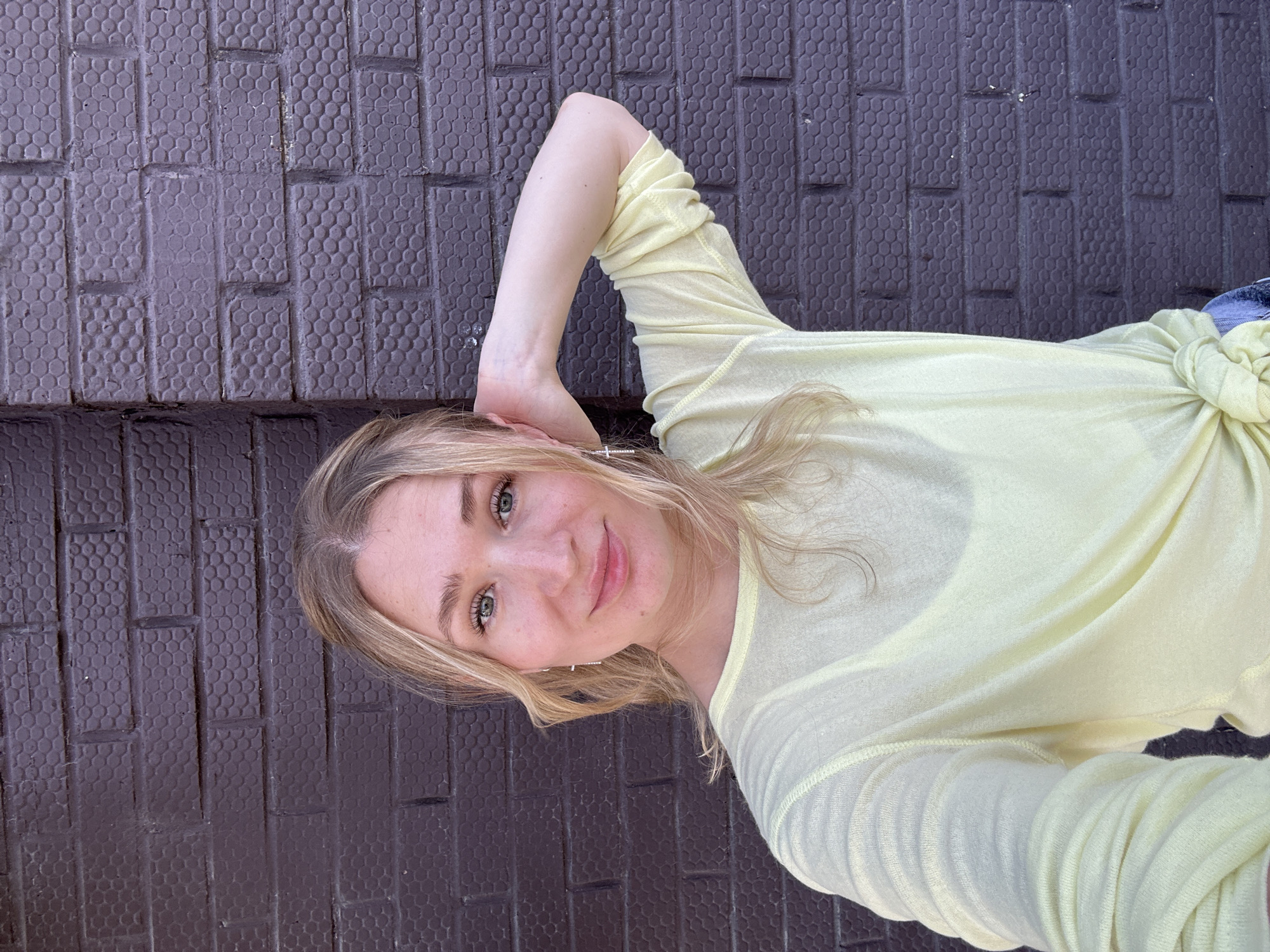 A young Caucasian woman with blonde hair is shown from the chest up, posing for a selfie in front of a dark purple brick wall. She is wearing a light yellow long-sleeved shirt that is tied at the bottom. Her right arm is bent at the elbow and her hand is resting on the back of her head. She is looking directly at the camera with a slight smile. The brick wall has a hexagonal pattern. The image does not depict the city of Verkh-Sosnova, Russia.