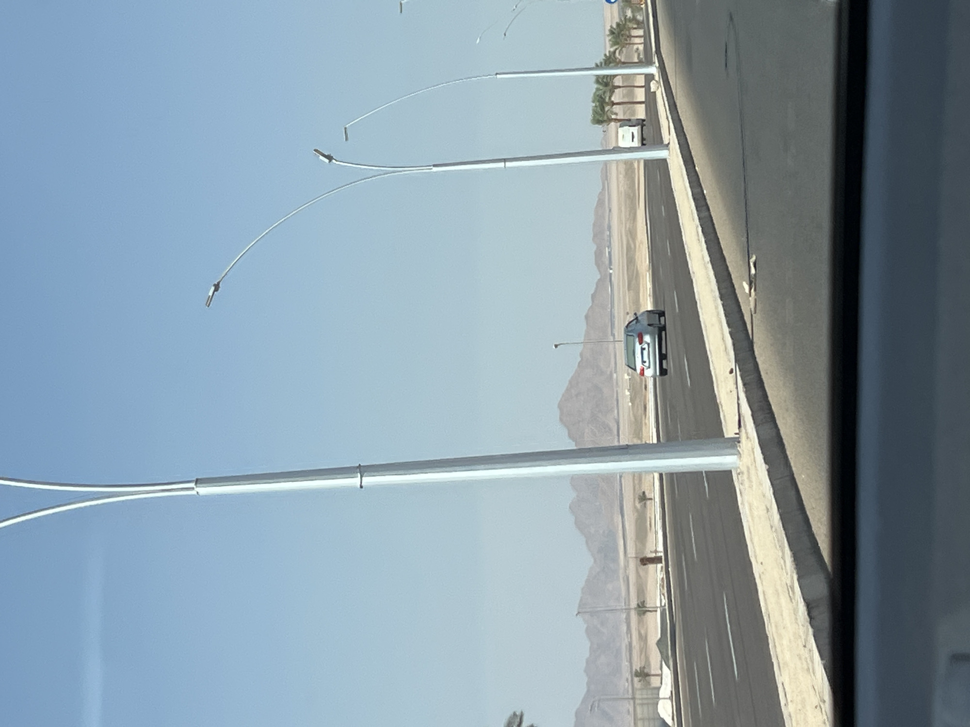 The image depicts a wide, paved road in Sharm El Sheikh, Egypt, with a clear blue sky overhead and arid, mountainous terrain in the distance. Several modern streetlights with curved arms line the right side of the road, spaced at intervals. A light-colored sedan is traveling in the left lane, heading away from the viewer. Further down the road, a white van and several palm trees are visible. The roadside features a concrete barrier and a patch of dry, sandy ground. The overall scene suggests a relatively new or developing area within the city, characterized by its infrastructure and natural desert landscape.
