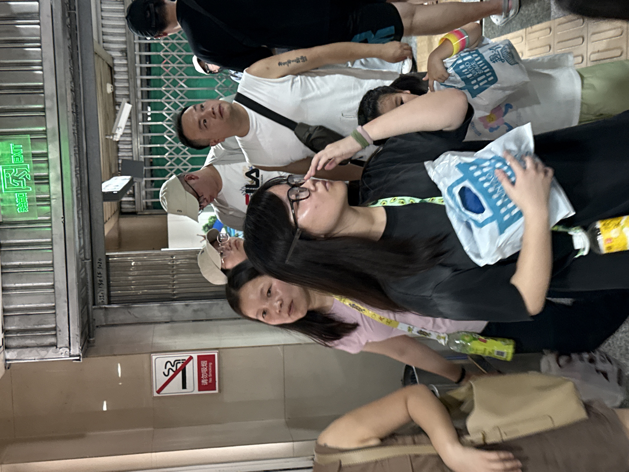 In Yuzhong District, China, a group of people are gathered near an entrance marked with a green "EXIT" sign. A "No Smoking" sign is visible on the wall to the left. Several individuals are holding shopping bags, some of which appear to be reusable grocery bags with blue basket designs. One person is wearing a white sleeveless shirt and has a black bag slung across their chest. Another individual, a young girl, is wearing a white shirt and colorful bracelets on her arm. The environment appears to be an indoor or covered public space with metallic gates and a tiled floor.