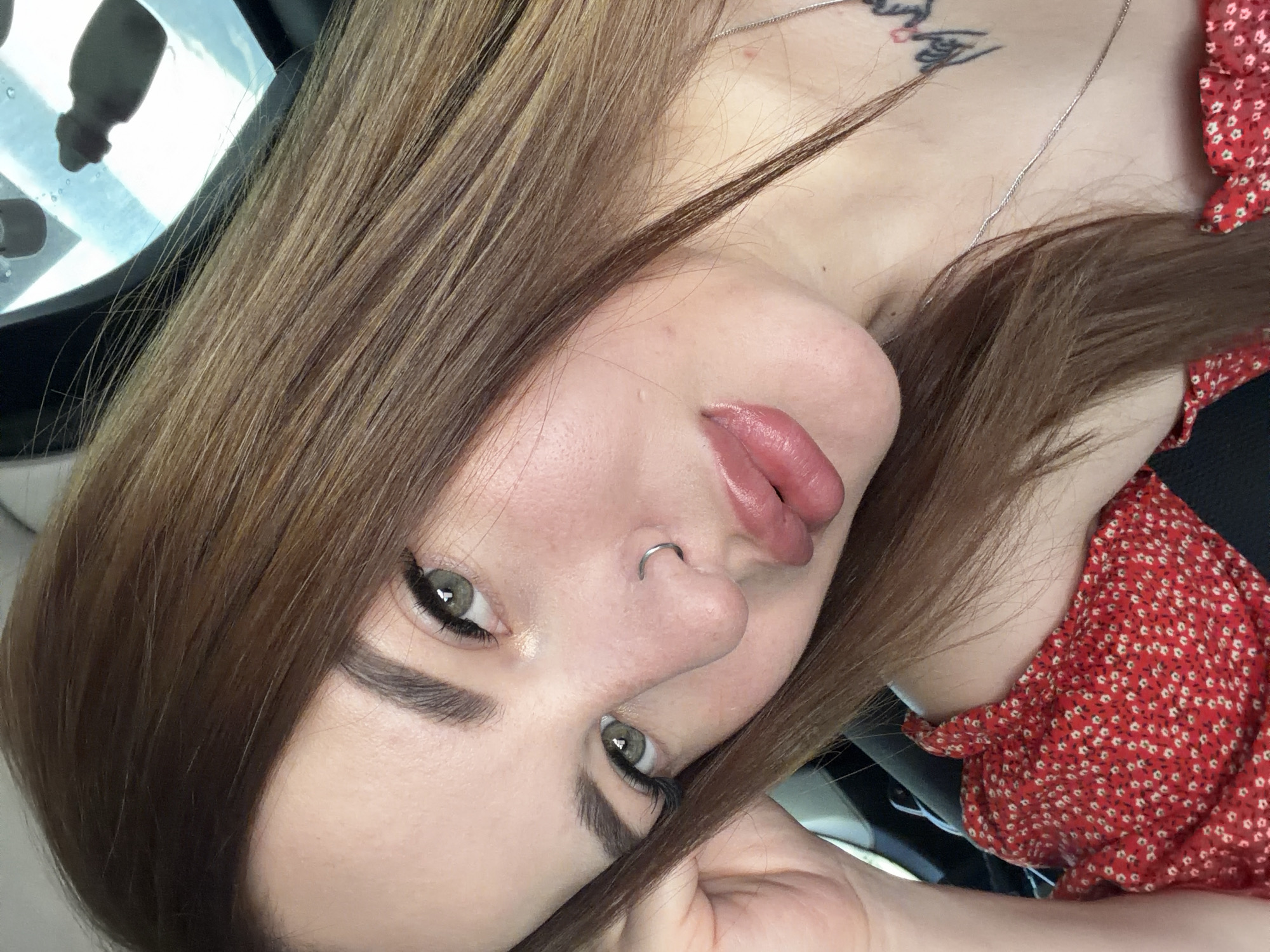 The image displays a female subject in an Unknown City, Unknown Country, captured in a medium close-up from the chest up, positioned inside what appears to be a vehicle. The subject has fair skin, medium-length brown hair, and green eyes. She exhibits well-defined eyebrows and prominent eyelashes. A silver hoop nose ring is present in her left nostril. Her lips are full and appear to be enhanced with a light pink or nude lip product. Small skin imperfections or pores are visible on her face. She is wearing a red top adorned with a small white floral pattern, featuring a wide or off-shoulder neckline. A thin silver chain necklace is visible around her neck, and a black tattoo with red accents is located on the right side of her upper chest/neck area. Her left hand is partially visible, resting near her left temple. The subject is looking directly into the camera with her head slightly tilted. The background in the upper right quadrant shows a blurred, bright light source with dark silhouettes, consistent with reflections or objects seen through a vehicle's window or sunroof, along with minor surface imperfections resembling water droplets.