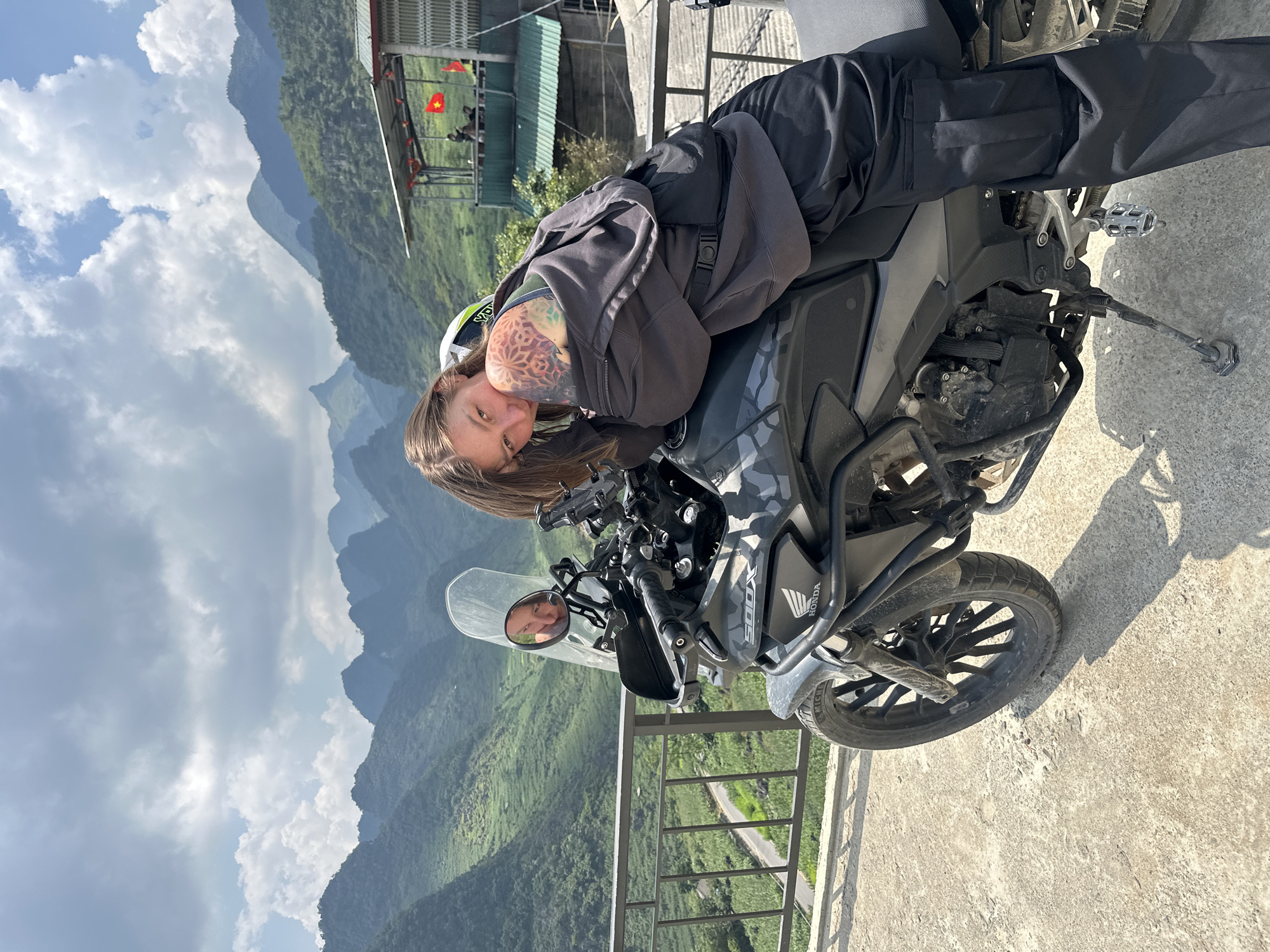 A woman with brown hair and a tattoo on her left arm is reclining on a gray and black Honda CB500X motorcycle. The motorcycle is parked on a concrete platform near a black metal railing overlooking a mountainous landscape. The location is Lũng Phìn Commune, Vietnam, as indicated by the scenery. Behind the motorcycle and railing, there are green mountains and a cloudy sky. The woman is wearing a gray hooded sweatshirt and black pants. In the motorcycle's windshield, a reflection of the woman's face can be seen. In the background, there is a structure with a green roof and a Vietnamese flag.