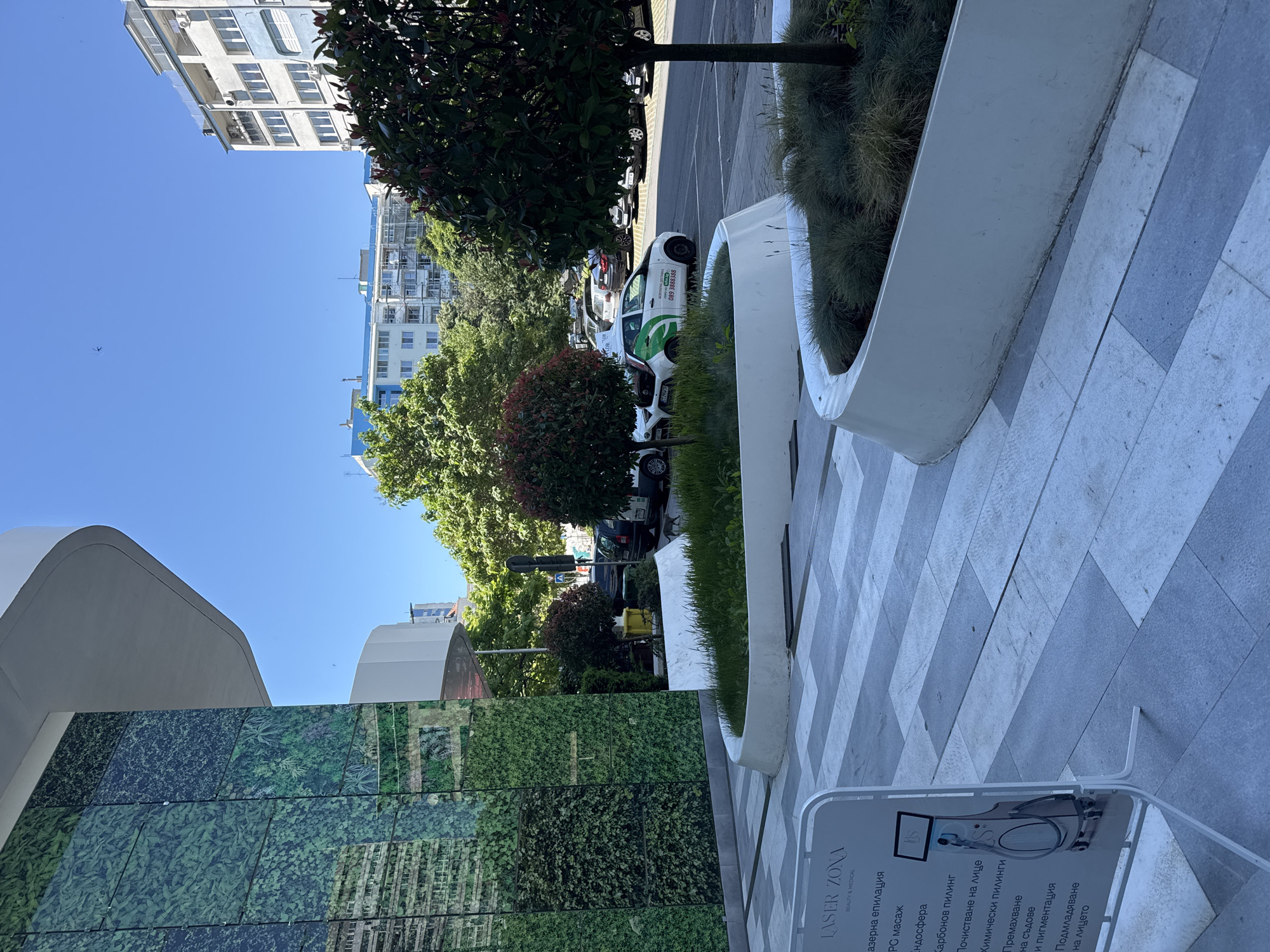 This is an eye-level, medium shot of a modern building facade and a pedestrian walkway in Burgas, Bulgaria. The building's left side features a large, green wall covered in what appears to be artificial foliage, reflecting the sky and surrounding buildings. To the right of the facade, a white, curved planter box is filled with grass and small plants. Beyond the planter, a paved road is visible, with several parked cars, including a white and green delivery van. Trees with red-tinged foliage are planted in front of the building. In the background, several multi-story residential buildings with balconies and air conditioning units are visible under a clear blue sky. A white sign with text in Cyrillic script, advertising "LASER ZONA BEAUTY & MEDICAL," is positioned on the left side of the walkway. The overall scene depicts an urban environment with elements of landscaping and commercial activity.
