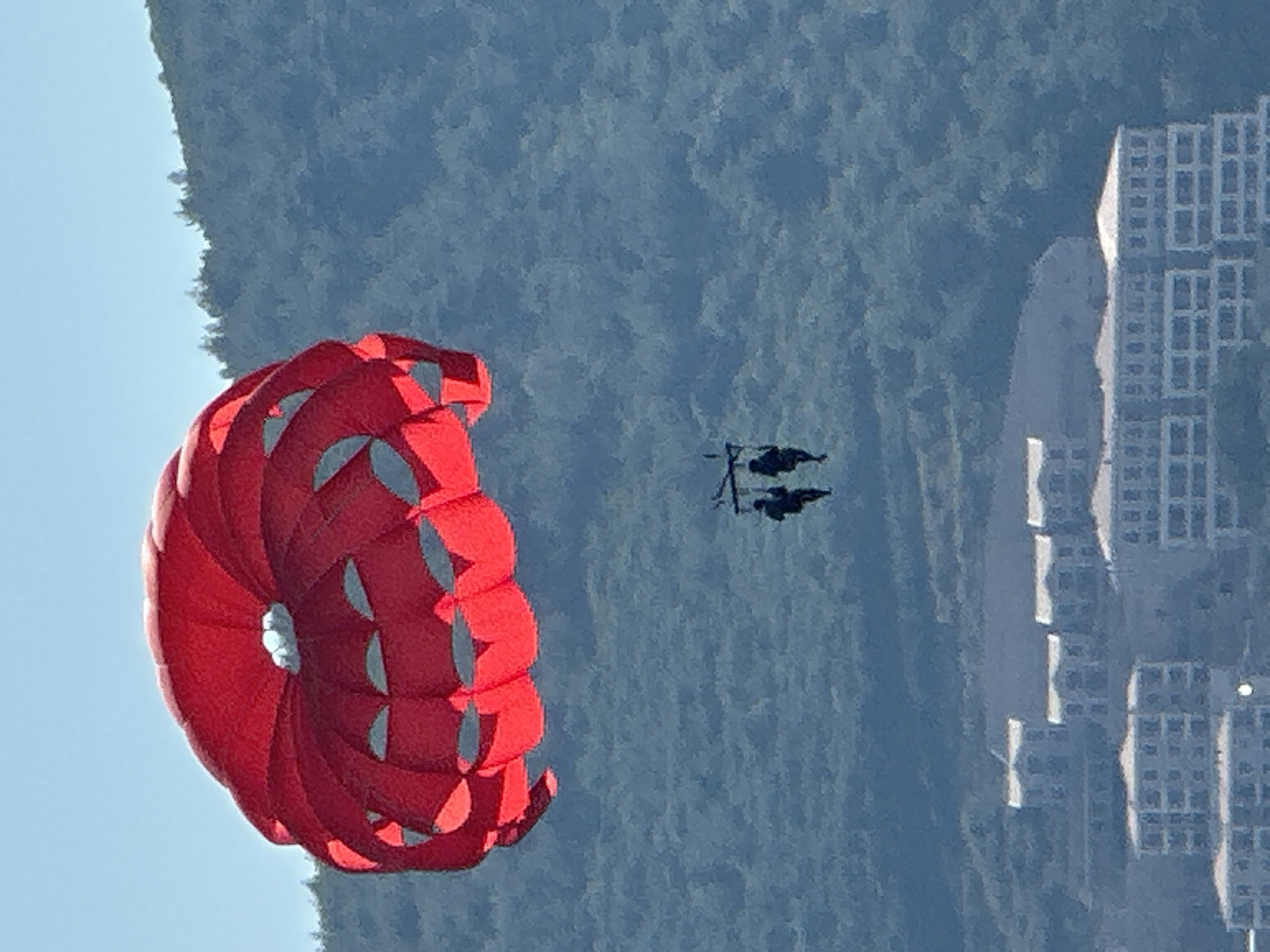 A bright red parasail, with a visible central opening and scalloped edges, is suspended in a clear blue sky. Below the parasail, two figures are harnessed and appear to be parasailing. The background is dominated by a lush, green, tree-covered mountainside, which slopes downwards towards a coastline. In the foreground, a cluster of white buildings with light-colored roofs are visible, suggesting a resort or residential area situated along the coast. This scene is consistent with the picturesque coastal scenery found in Budva, Montenegro.