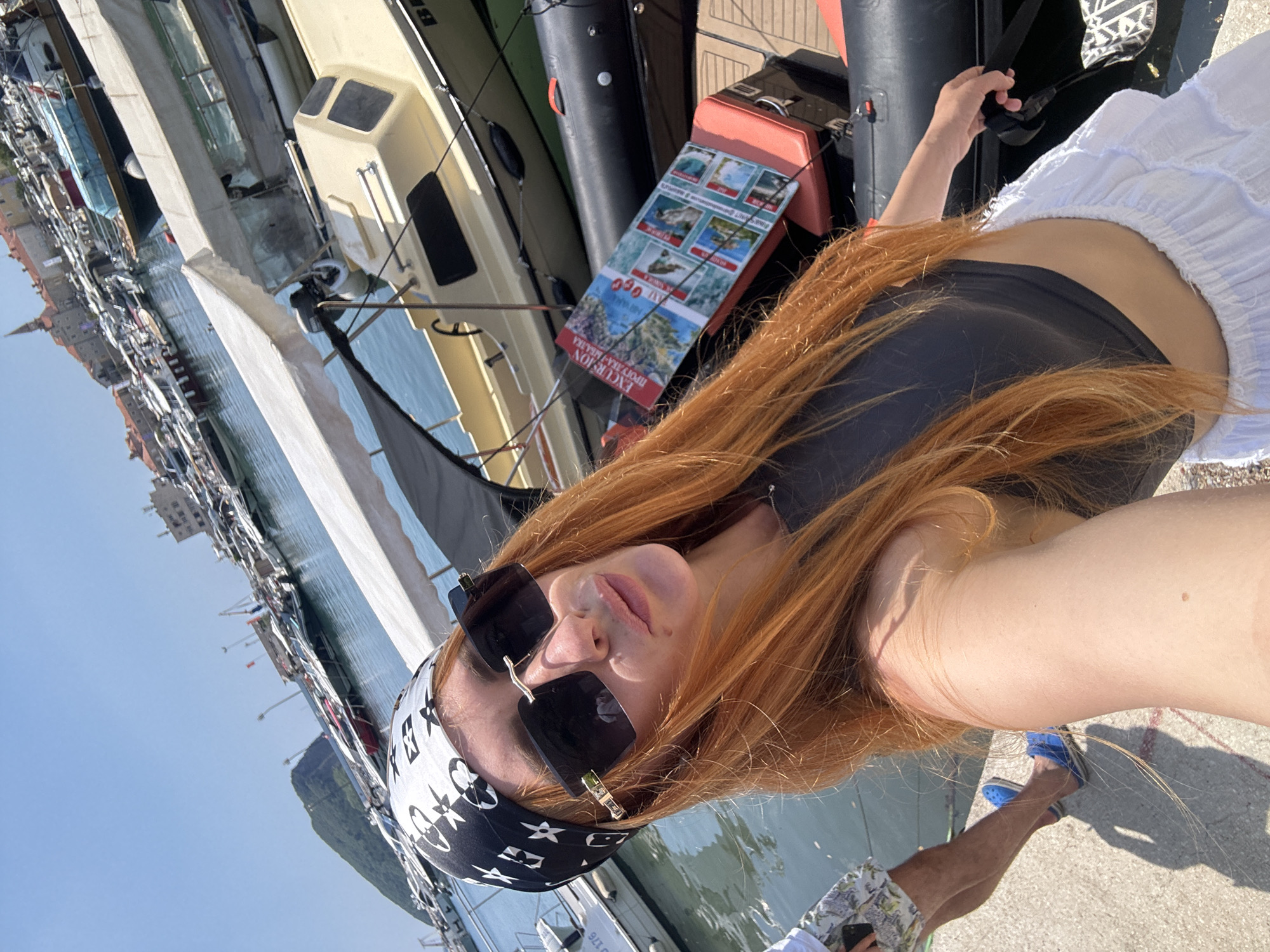 This media file shows a woman with long, red hair and a bandana on her head. She is wearing sunglasses and a black top, and her right arm is visible. She is leaning against a boat or a dock. In the background, there are several boats docked in a harbor, and buildings are visible in the distance. The water is calm, and the sky is clear and blue. The image appears to be taken on a sunny day. There is text on a brochure visible in the image that reads "EXCURSION PROVRAKA-MLAKVA". The overall scene suggests a leisure activity, possibly a boat tour or a day at the harbor. The location context provided is Budva, Montenegro.