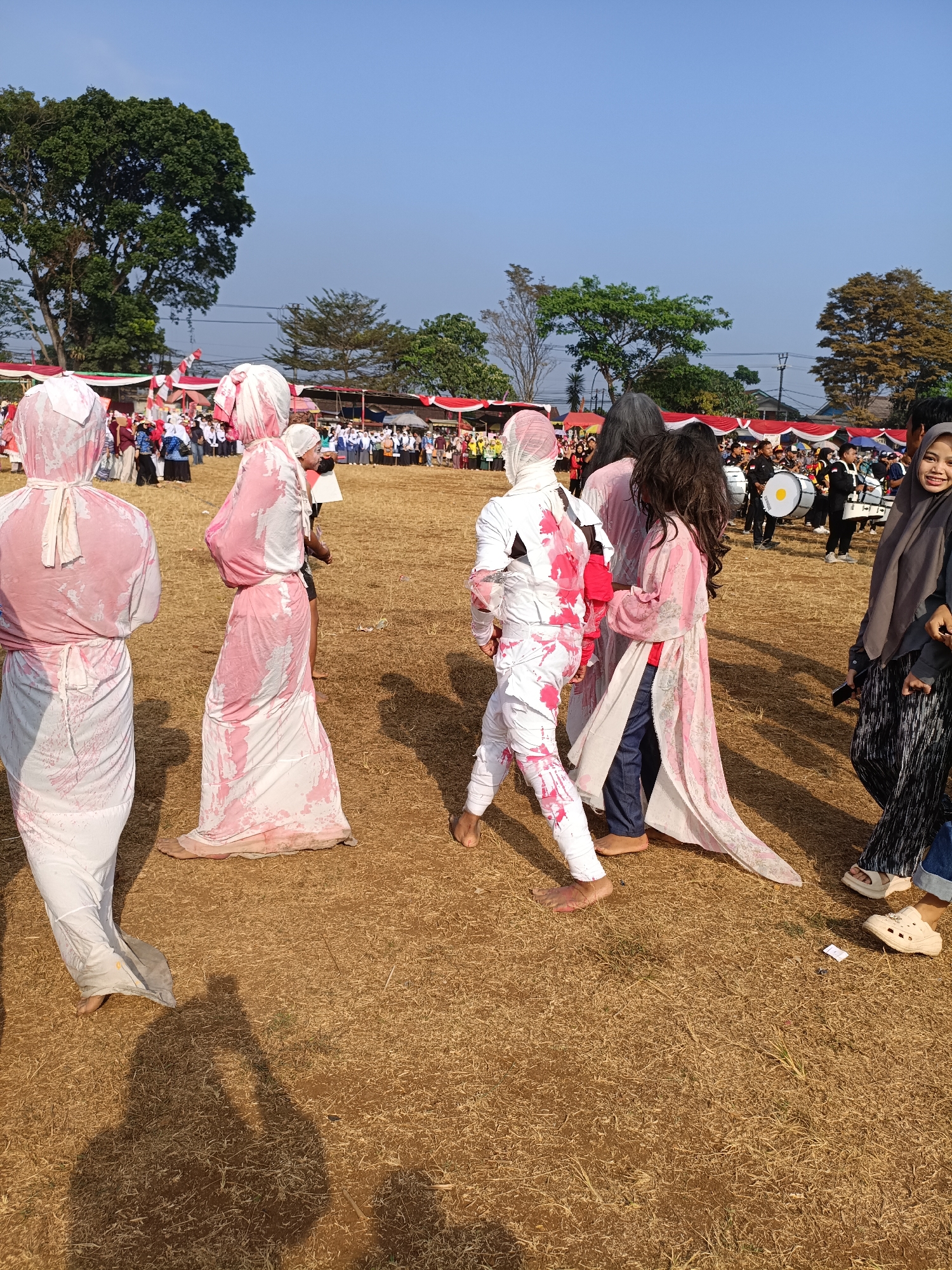 In Cibodas, Indonesia, on a dry, grassy field under a clear blue sky, a group of individuals are gathered, possibly for a parade or performance. Several people in the foreground are wearing white outfits with pink splatters, resembling a paint or dye. These outfits are long and flowing, some covering their heads. One person in the center wears a white outfit with more pronounced red splatters. To the right, a woman in a hijab smiles at the camera. In the background, a band with drums is visible, with tents and crowds of people under a bright sun. The overall scene suggests an outdoor event with participants in costume.