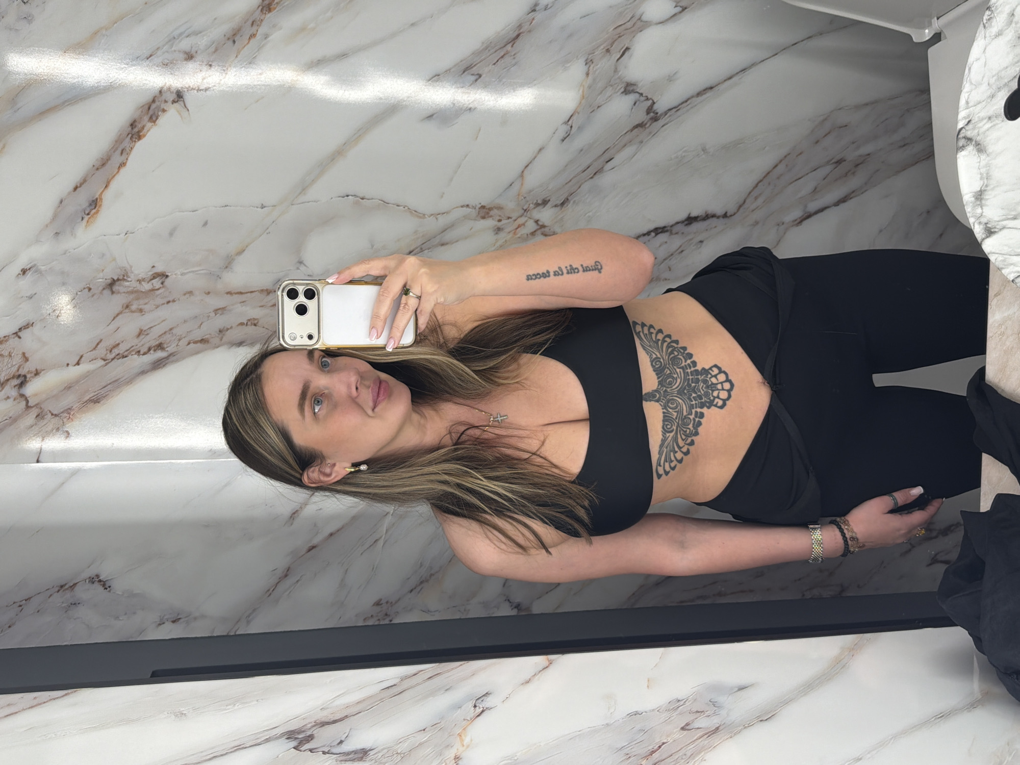 A woman is captured in a mirror selfie in what appears to be a bathroom in Moscow, Russia. She is holding a white smartphone with multiple cameras in her left hand, which is angled upwards. The woman has long, light brown hair, and is looking towards the phone's camera with a neutral expression. She is wearing a black sports bra or crop top, revealing a large, intricate dark tattoo on her right rib cage/abdomen, and black leggings.
Her left forearm bears a tattooed English phrase, which, when un-mirrored, reads "just do it all." On her left ring finger, she wears a ring, and a cross pendant hangs from a necklace around her neck. Her right wrist is adorned with multiple bracelets, including a metallic one and several beaded ones. The background features polished walls and surfaces with a distinctive white and gray/brown marble pattern. A white toilet, also covered in a marble pattern, is visible in the upper right corner of the frame. The setting appears modern and well-lit.