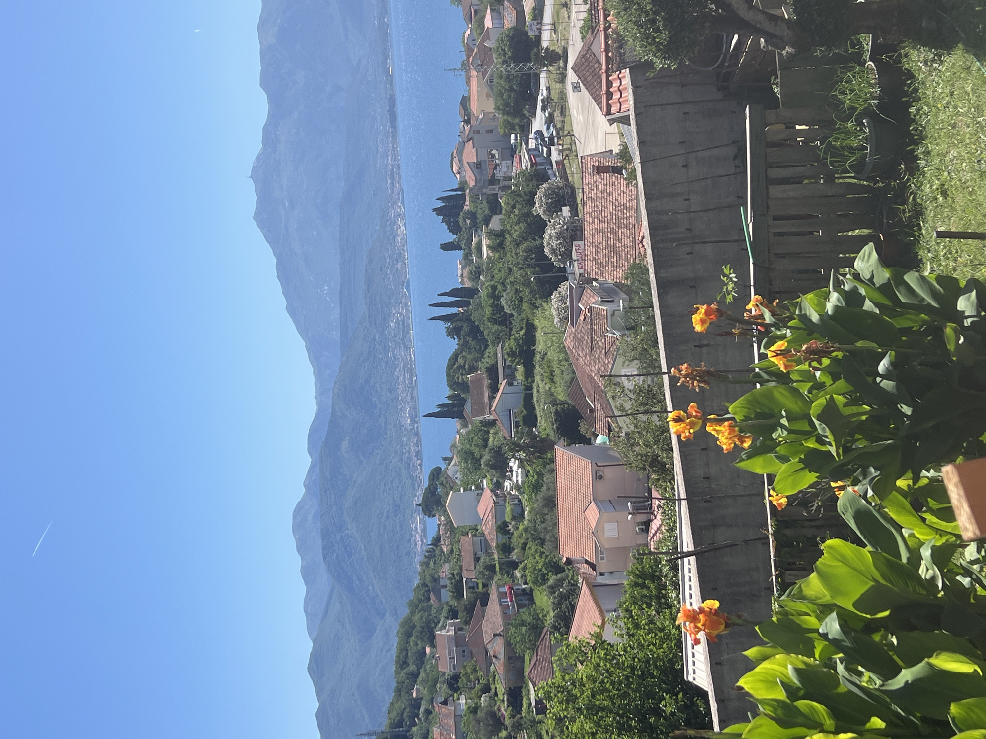 The image presents a scenic view of Baošići, Montenegro, featuring a hillside residential area overlooking a body of water. Mountains are visible in the distance under a clear blue sky. The foreground includes foliage, specifically green plants with orange flowers. A concrete and wooden fence-like structure is present, likely part of a property boundary. Buildings with red-tiled roofs are interspersed among trees and vegetation. A contrail is visible in the sky. The image is taken at an angle, tilting slightly to the left.