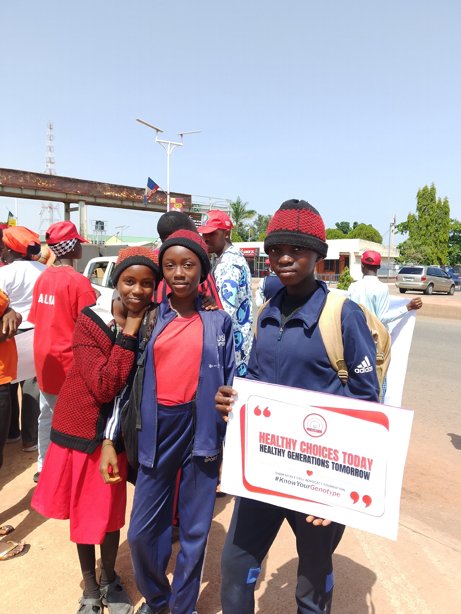 In Jos, Nigeria, a group of people stand on a dirt road, some holding a sign that reads "HEALTHY CHOICES TODAY HEALTHY GENERATIONS TOMORROW" and "#KnowYourGenotype". Three young women are prominently featured in the foreground, two of whom are looking at the camera. The woman on the far left wears a red knitted sweater over a red dress and black shoes. The woman in the center wears a red t-shirt and a blue athletic jacket with a backpack. The woman on the right wears a blue athletic jacket and pants, also with a backpack. Other individuals in the background are wearing red caps and various colored shirts. The environment appears to be a street with buildings and trees in the distance, under a bright, clear sky.