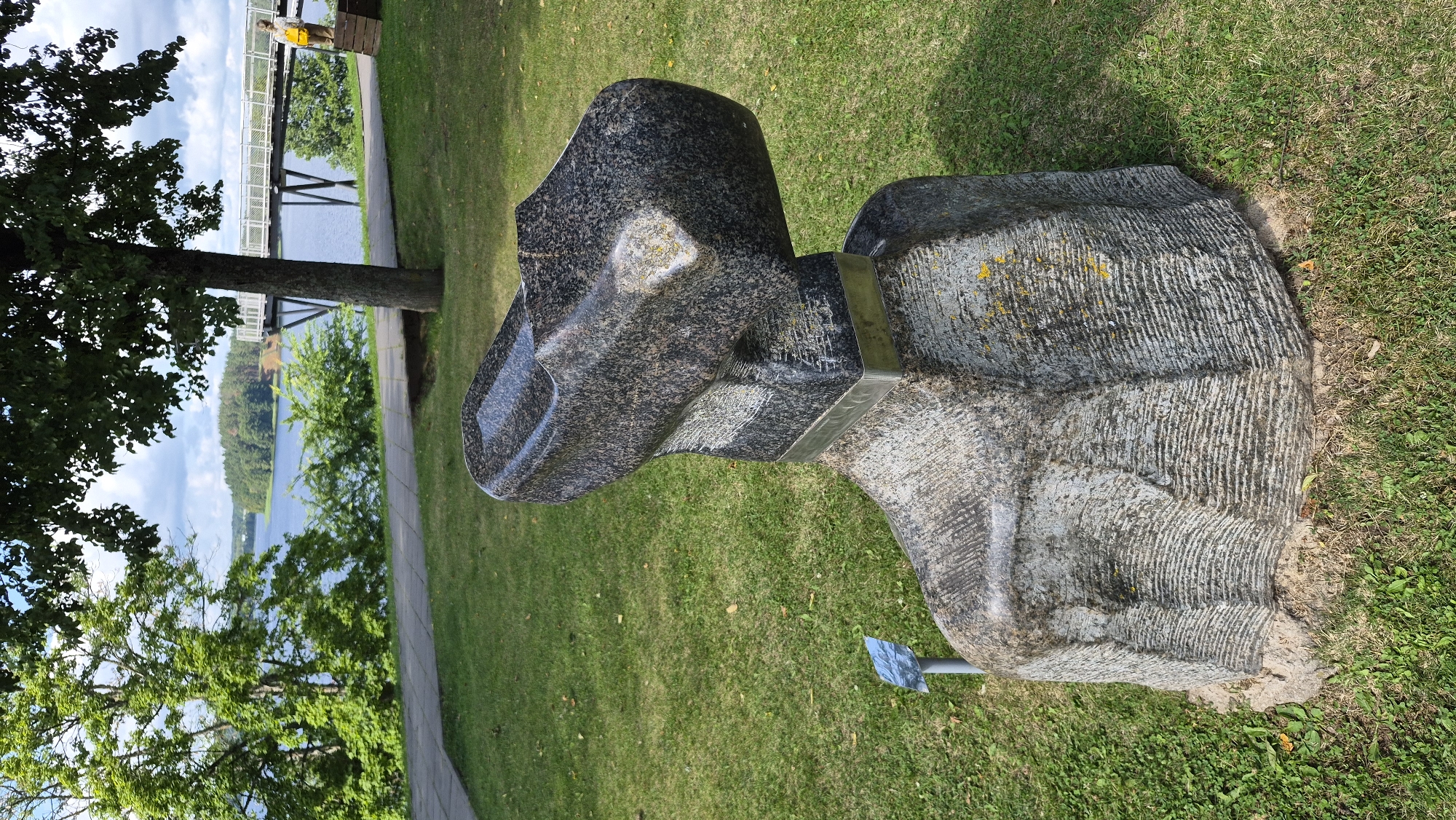 The image shows a stone sculpture shaped like a large clog, placed outdoors on a grassy area. The clog is made of dark granite with a textured, rough base. A silver band encircles the clog where its upper part meets the base. In the background, there is a body of water, likely a lake, with trees and a pedestrian walkway. A white railing structure and a person wearing a yellow backpack are visible on the right side of the background. The sculpture is located in the city of Zarasai, Lithuania, specifically near the Zarasai Lake promenade.