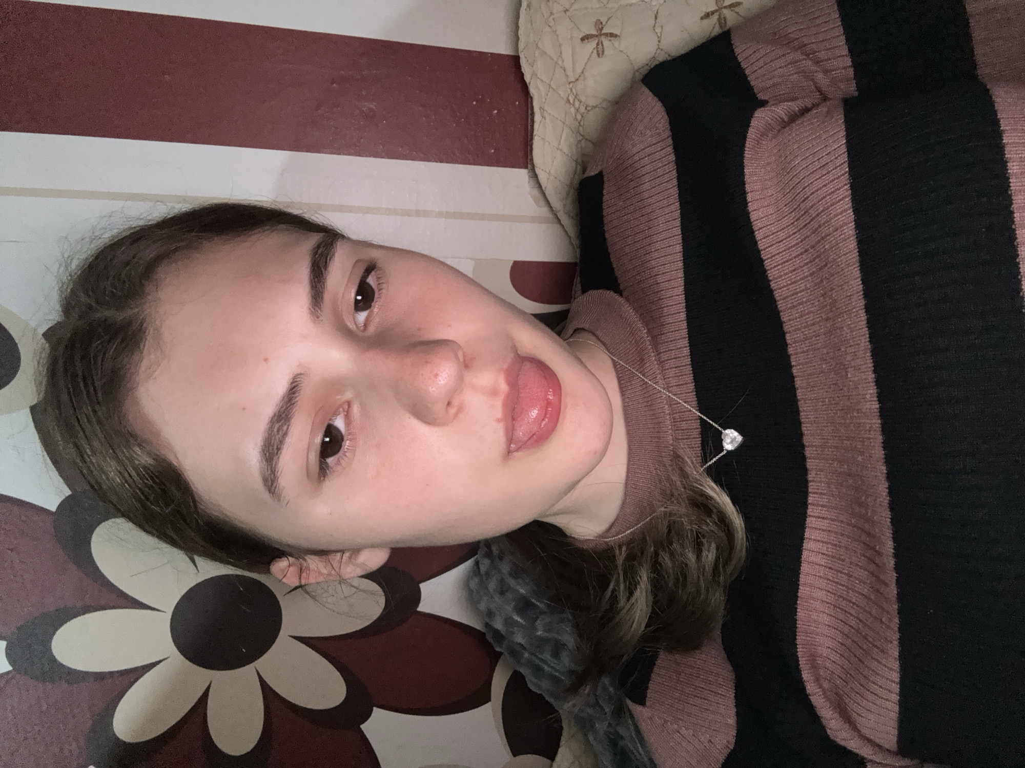 The image captures a close-up of a young woman lying down in an indoor setting. She has dark hair, dark eyebrows, and dark eyes, looking directly at the camera with a playful expression, her tongue slightly extended. Her complexion is fair, and she appears to have minimal or no makeup. She is wearing a ribbed sweater with wide horizontal stripes in a muted pinkish-brown (or mauve) and black. A delicate silver chain necklace with a small, heart-shaped, faceted pendant rests on her chest.
The background consists of several distinct patterns and textures. Below her, a flat surface, possibly a rug or bedspread, displays a repeating floral pattern featuring large, stylized blossoms in dark brown, off-white, and black. Above and behind her head, a light-colored, possibly cream, quilted surface with an embroidered cross-like design is visible. Near her neck and shoulder, a soft, grey, furry material, likely a blanket or pillow, adds another texture to the scene.
The lighting suggests an indoor environment, without direct cues for time of day, though it appears to be artificial light. No specific outdoor weather conditions are discernible. The overall mood is casual and relaxed, with a hint of playfulness conveyed by her expression. There is no visible text in the image. While the image itself does not contain explicit geographical cues, the provided context indicates the location is Panagyurishte, Bulgaria.