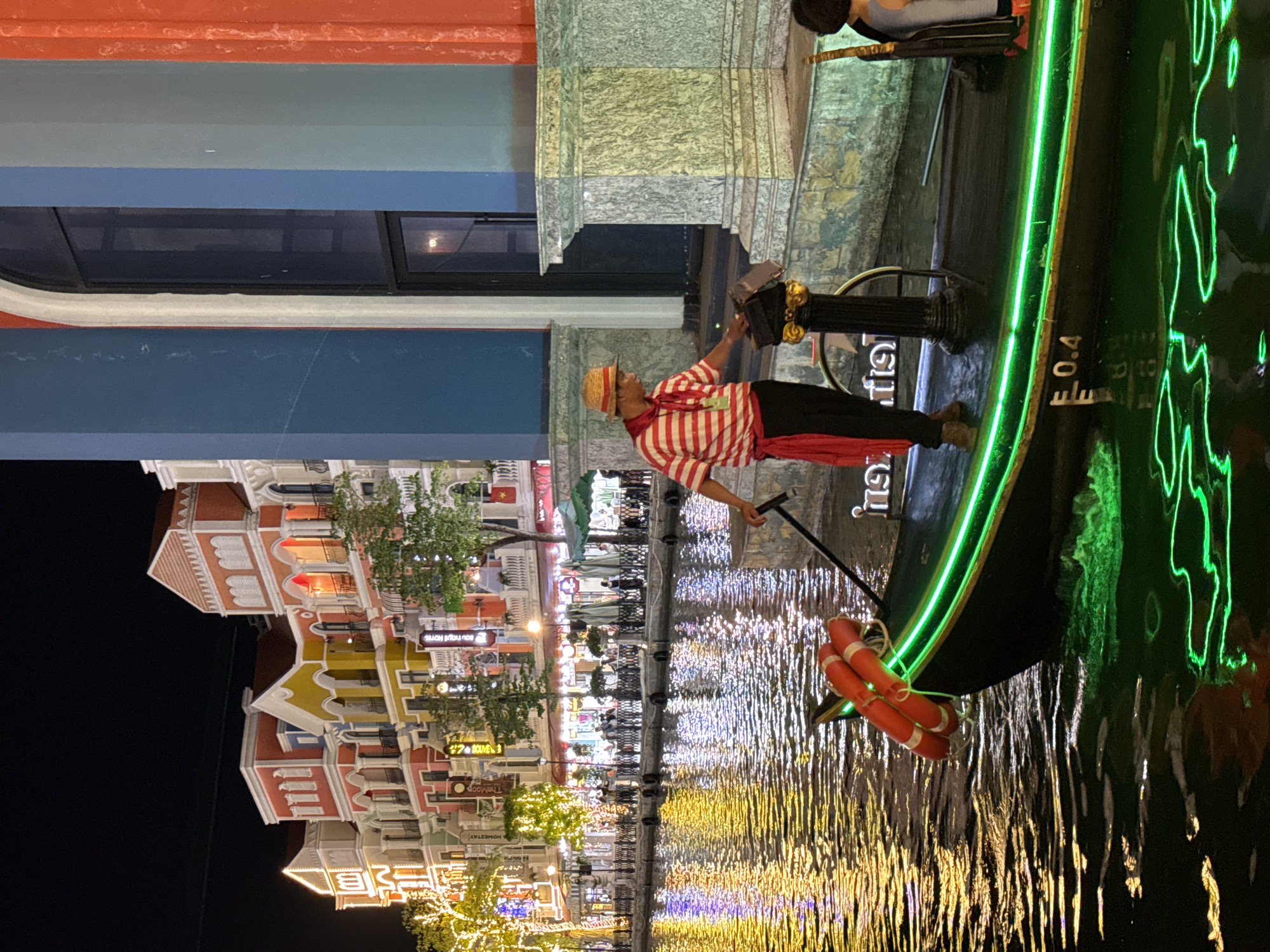 The image displays a vibrant night scene along a canal, likely a themed attraction resembling Venice, located in Phú Quốc, Vietnam. A person, dressed as a gondolier in a red and white striped shirt, red scarf, and straw hat, stands in a dark gondola, holding a long pole used for propulsion. Another person is partially visible seated towards the back of the boat.
The gondola is distinctly outlined with bright green neon lighting, which creates striking reflections on the water. An orange lifebuoy is attached to the side of the boat, and the marking "E 0.4" is visible on its hull.
The canal is flanked by colorful, multi-story buildings featuring Venetian-style architecture with arches, balconies, and varied rooflines. These structures are extensively illuminated with warm decorative string lights and other fixtures, creating a lively and festive atmosphere. The abundant artificial lights from the buildings and the boat are brightly reflected on the dark water, forming shimmering patterns of yellow, white, blue, and green. Trees adorned with lights are also visible along the canal bank.
Visible text on various signs includes "BOUTIQUE HOTEL," "HOMESTAY," and "The LOOP." On a structure near the gondolier, Thai script can be seen, which reads "บริการ" (service). The dark sky confirms the scene takes place at night, and the clear reflections indicate calm water conditions. The overall impression is one of a picturesque and engaging tourist destination.