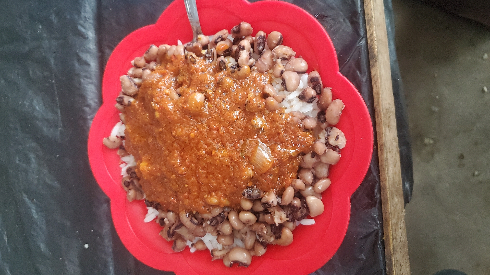 A close-up, top-down shot displays a red, flower-shaped plate filled with food. The plate contains a bed of white rice topped with black-eyed peas and a thick, reddish-brown sauce. The sauce appears to be a stew or a similar preparation, with visible chunks of onion and other ingredients. A silver spoon rests on the right edge of the plate, partially submerged in the food. The plate is set on a dark, textured surface, likely a table covered with a black plastic or oilcloth material. A wooden edge is visible at the bottom of the frame. The lighting suggests an outdoor or semi-outdoor setting, possibly a street food stall. While the image is set in Jalingo, Nigeria, the specific location within the city cannot be determined from the visual information provided. There are no people visible in the frame.