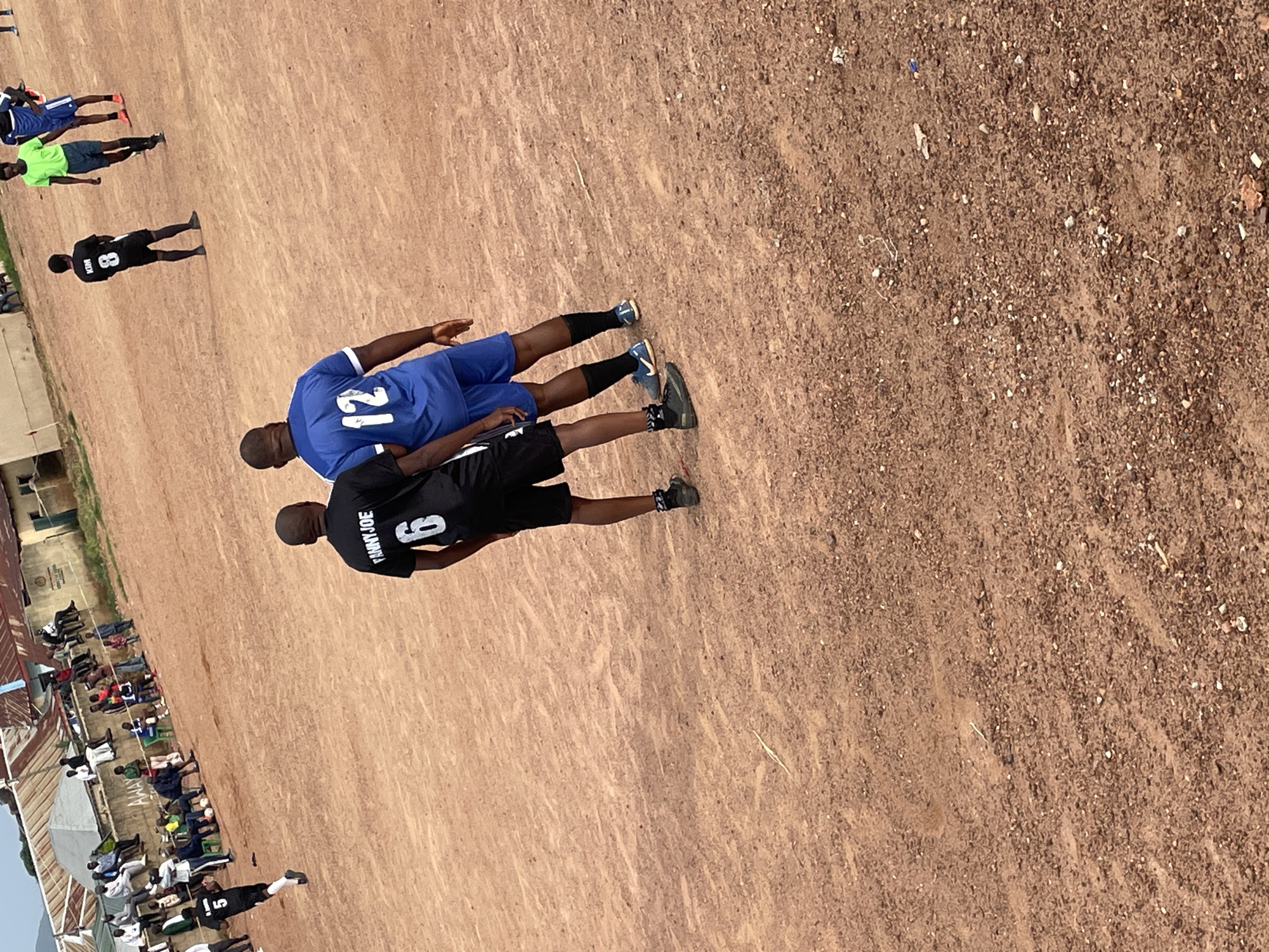 A medium shot captures two men in athletic attire standing on a dusty, red-brown outdoor field. The man on the left wears a black jersey with white lettering that reads "FANNY JOE" above the number "6," and black shorts. He has dark skin and short hair. The man on the right sports a bright blue jersey with the number "12" in white, and blue shorts. He also has dark skin and short hair. Both men are facing away from the camera and appear to be engaged in conversation or observing something beyond the frame.
In the background, several other individuals are visible, some seated and others standing, suggesting a sporting event or gathering. A few buildings with corrugated metal roofs are situated in the far distance, typical of informal settlements. The ground is uneven and littered with small stones and debris. The overall impression is of a casual outdoor activity in a somewhat arid, urban environment. The presence of what appears to be a soccer field and players suggests this location is within the city of Jos, Nigeria, known for its dusty, reddish soil and informal sporting facilities.