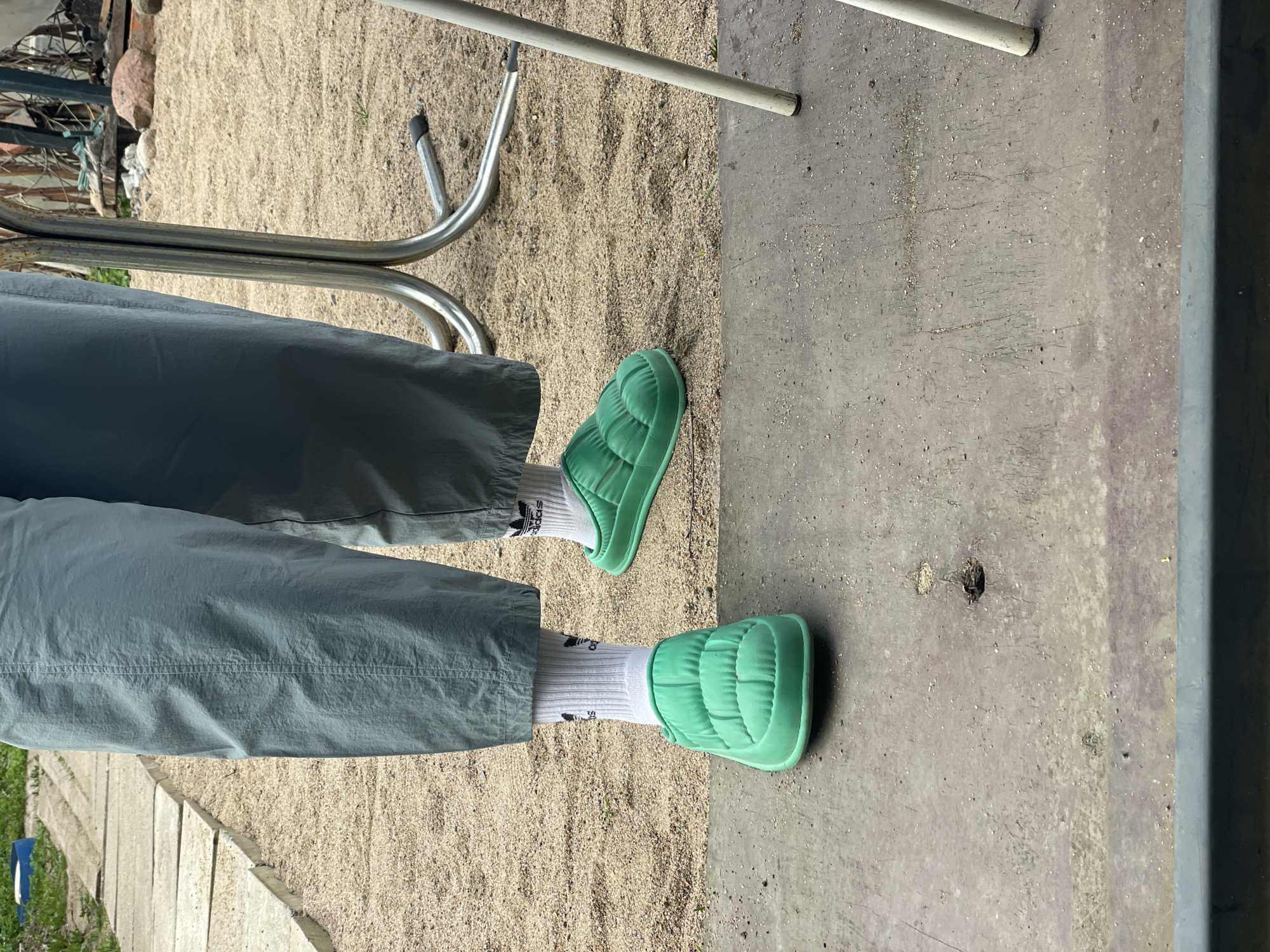 A person wearing green quilted slip-on shoes and white Adidas socks stands on an outdoor surface in what appears to be a backyard or patio. The person is wearing light green pants. The ground is partly covered in light-colored sand and partly in concrete. A metal chair is visible in the upper left of the image. The image was taken during the day. Location is inferred to be in Sestroretsk, Russia.
