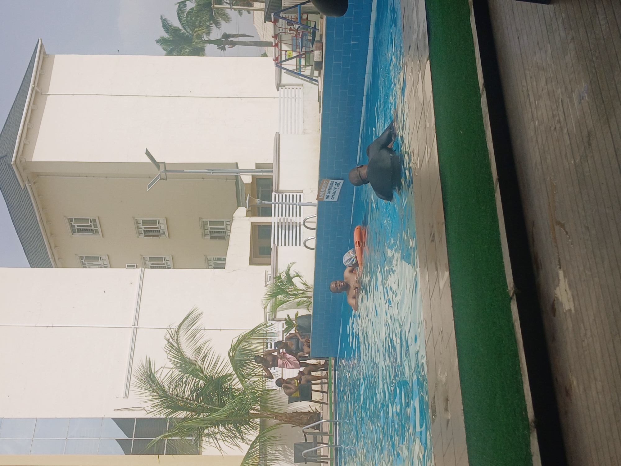 A group of people is at a pool next to a white building, most likely a hotel or residence. In the pool, one person is in a wetsuit, and another is holding a flotation device. On the deck of the pool, under a palm tree, are several people sitting. The pool is surrounded by a strip of artificial green turf. A sign reads "Warning: No Lifeguard on Duty". The location is Barakin Lerawi, Nigeria.