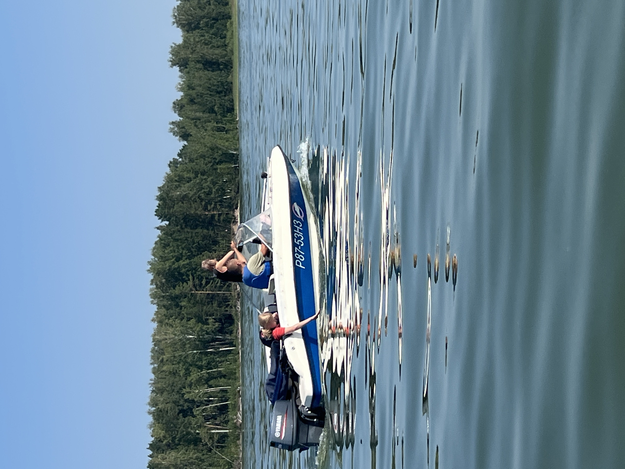 The image depicts a small, white and blue motorboat with the registration number P87-53H3 on a body of water. The boat has a Yamaha outboard motor. A person wearing a black top and a blue life vest is steering the boat. Another person, a child wearing a red shirt, is leaning over the side of the boat. A dark bag is also visible in the boat. The background shows a dense forest along the shoreline. The water is calm with visible ripples and reflections. The sky is clear and blue. The location appears to be a lake in a natural setting, consistent with areas like Borovoe, Russia, known for its lakes and forests.