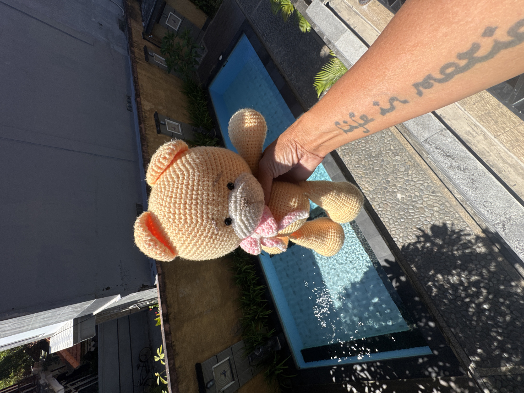 A person is holding a crocheted bear near a swimming pool in what appears to be Kuta, Indonesia, based on the architectural style and vegetation. The pool is rectangular and filled with water. A tattoo is visible on the person's arm, featuring text. The surroundings include buildings, paving stones, and shadows of trees, creating a sunlit environment.